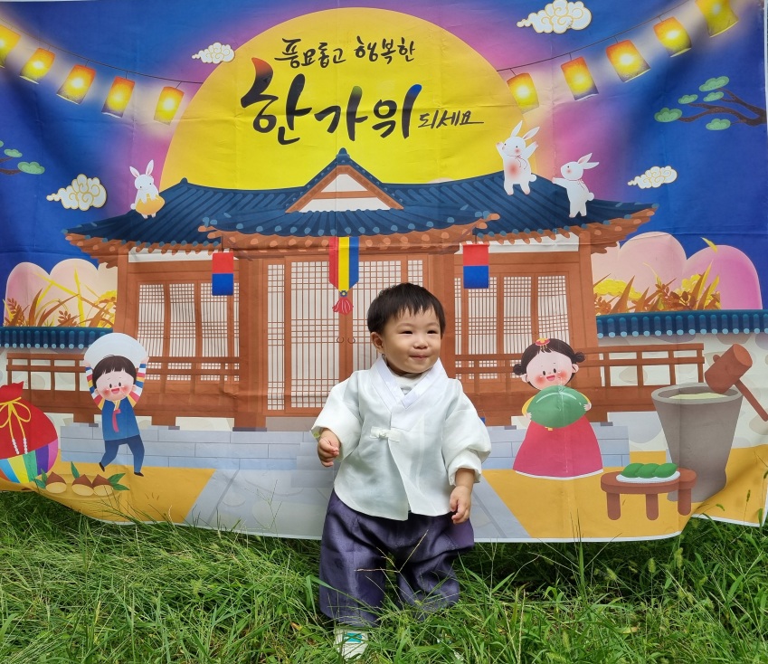 전통 한복을 곱게 차려입은 지호가 "풍요롭고 행복한 한가위 되세요"라고 쓰인 한옥 배경의 현수막 앞에 잔디밭에 서서 미소 짓고 있다.