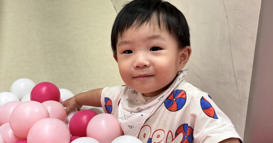 밝은 미소를 지으며 핑크색과 흰색 공이 가득한 볼풀장에 앉아있는 아기 지호의 모습. 베이지색 티셔츠와 반다나를 착용하고 있다.