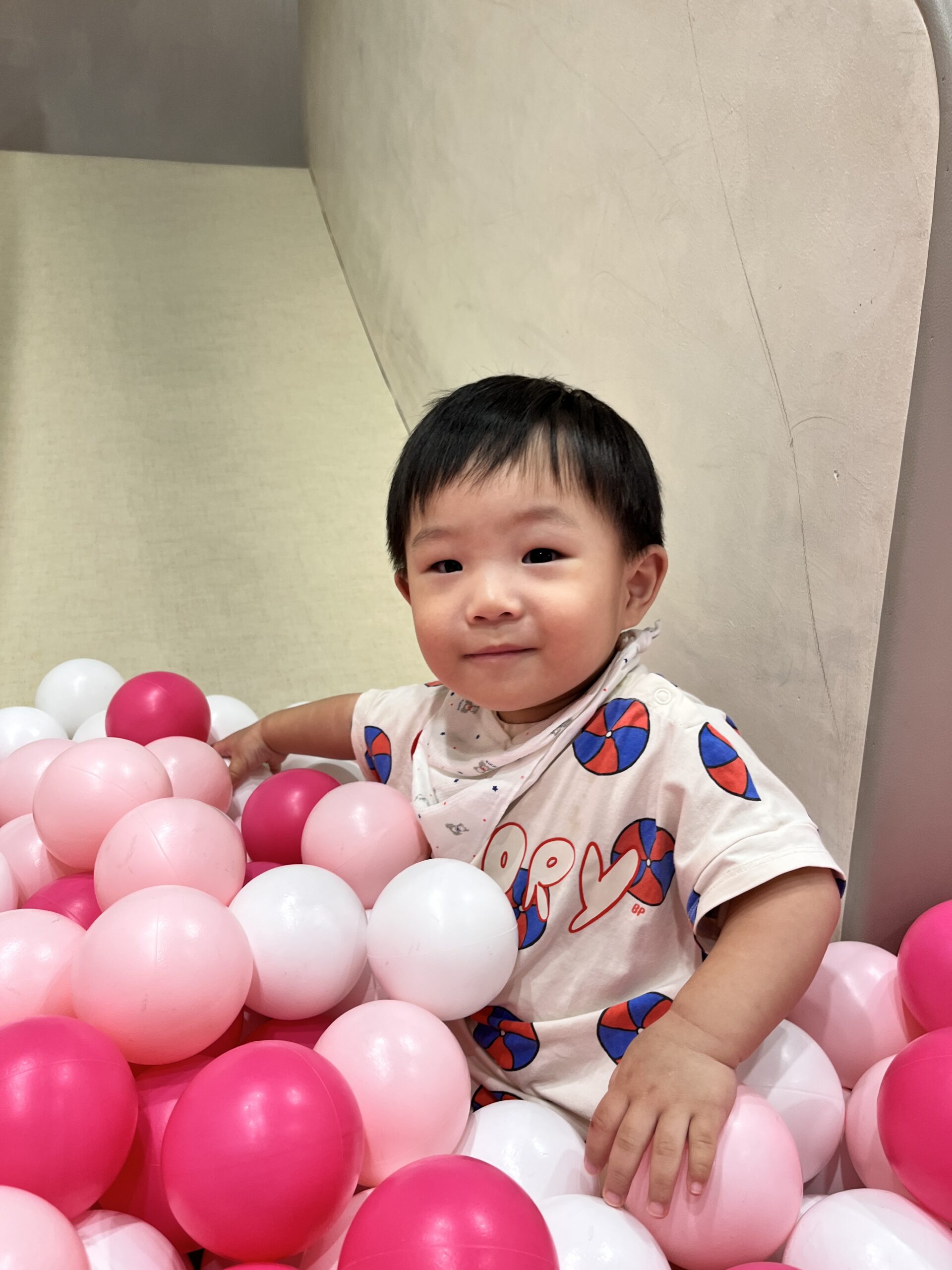 밝은 미소를 지으며 핑크색과 흰색 공이 가득한 볼풀장에 앉아있는 아기 지호의 모습. 베이지색 티셔츠와 반다나를 착용하고 있다.