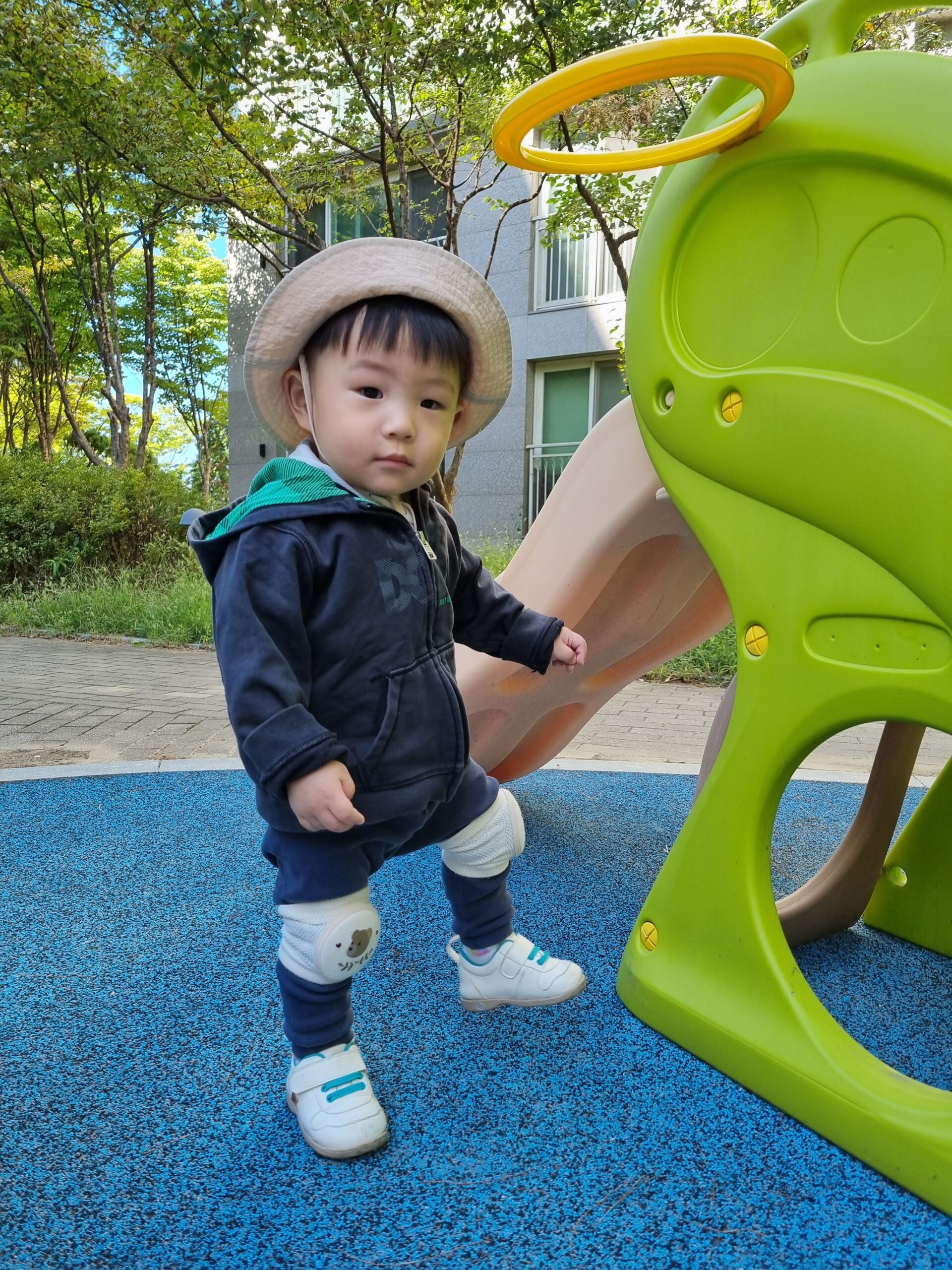 베이지색 모자와 후드티, 무릎 보호대를 착용한 아기 지호가 파란색 놀이터 바닥에 서서 미끄럼틀 옆에서 카메라를 진지하게 응시하고 있다. 뒤로는 나무와 아파트 건물이 보인다.