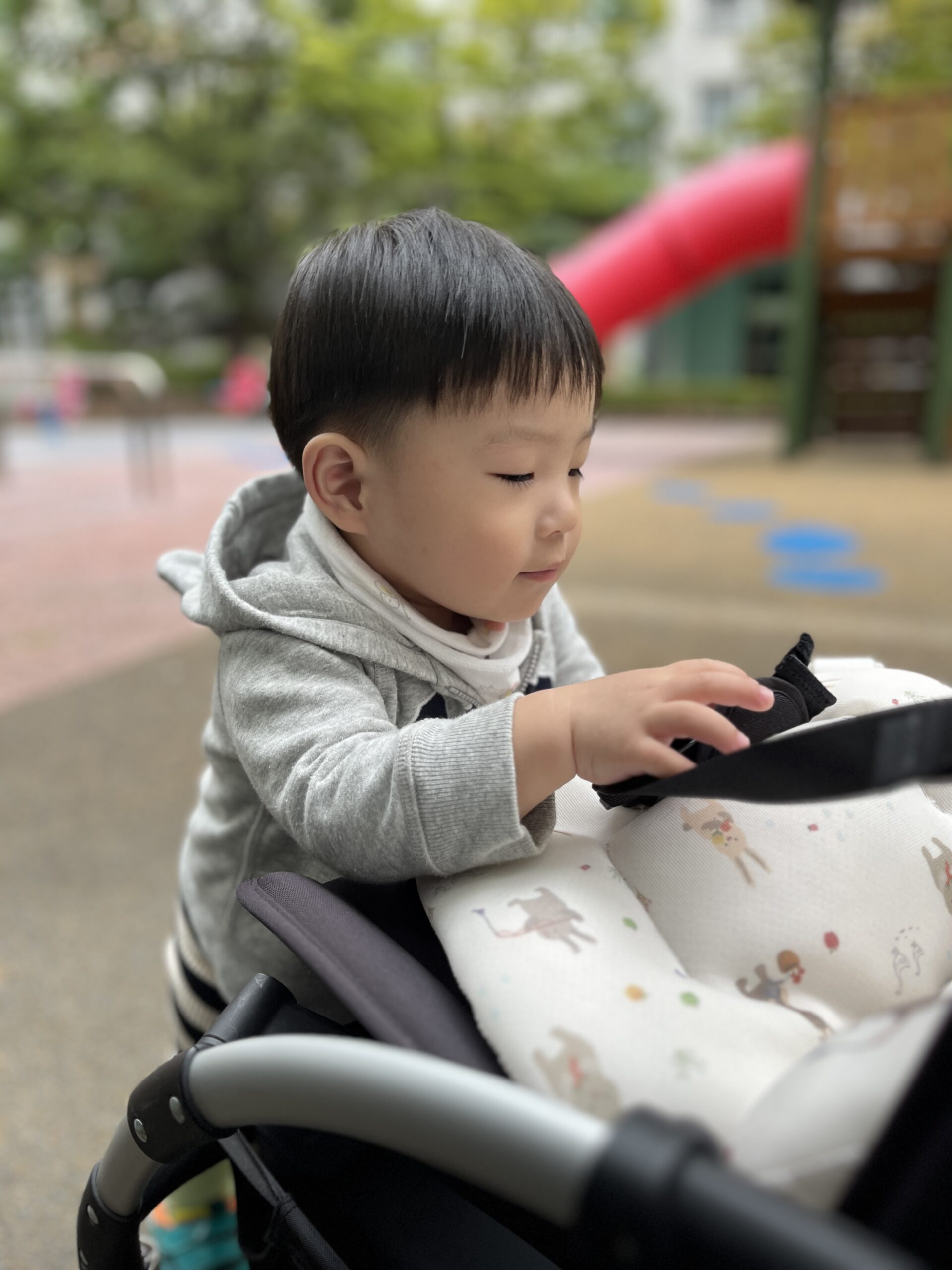 회색 후드티를 입은 지호가 야외 공원 같은 곳에서 유모차에 앉아 아래를 내려다보며 멜빵을 만지고 있다. 뒤로는 흐릿하게 붉은색 미끄럼틀이 보인다.