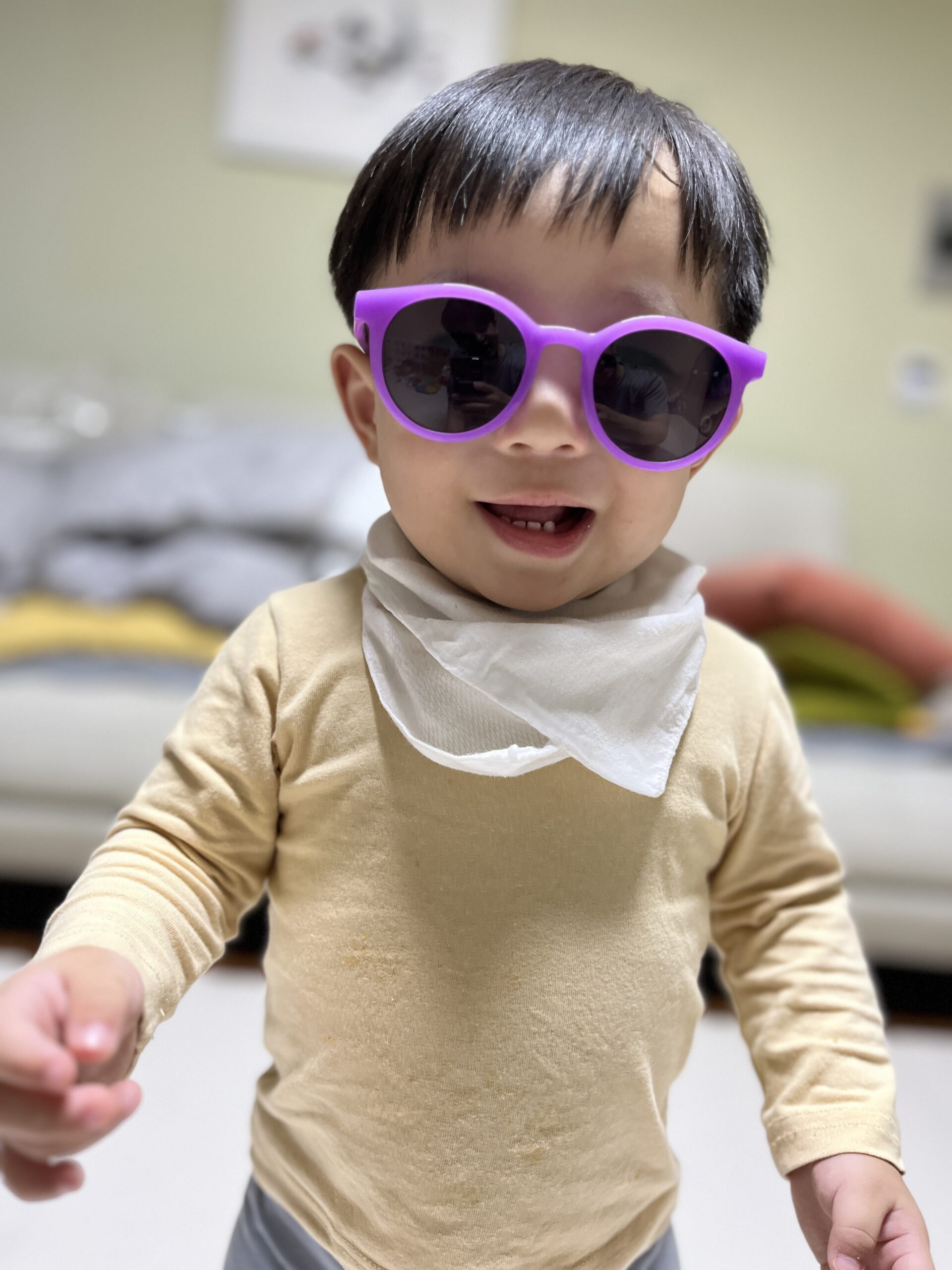 보라색 선글라스를 끼고 환하게 웃고 있는 아기 지호의 상반신 클로즈업 사진. 베이지색 상의와 흰색 턱받이를 하고 있으며, 오른손을 앞으로 뻗고 있다.