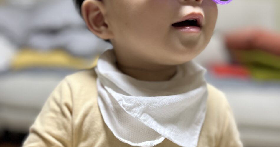 실내에서 베이지색 긴팔 티셔츠와 흰색 턱받이를 한 아기가 보라색 선글라스를 쓰고 비스듬히 위쪽을 바라보고 있다. 입이 살짝 벌어져 있고 표정은 다소 진지하며, 배경은 흐릿한 실내 공간이다.