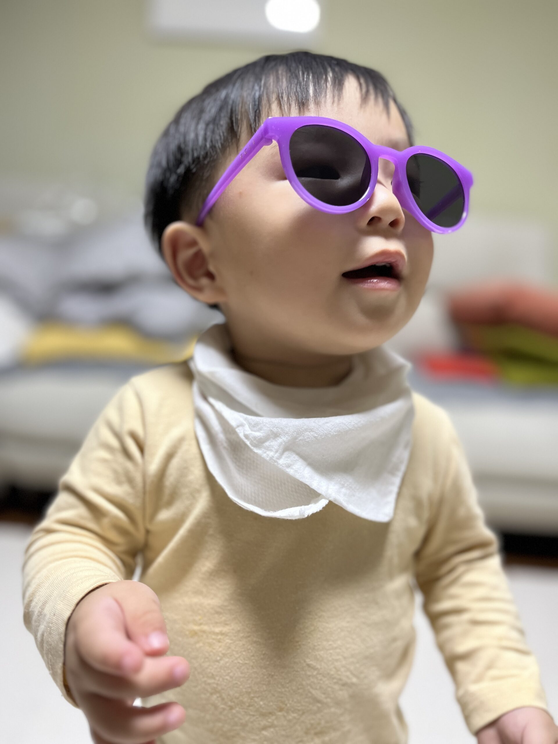실내에서 베이지색 긴팔 티셔츠와 흰색 턱받이를 한 아기가 보라색 선글라스를 쓰고 비스듬히 위쪽을 바라보고 있다. 입이 살짝 벌어져 있고 표정은 다소 진지하며, 배경은 흐릿한 실내 공간이다.