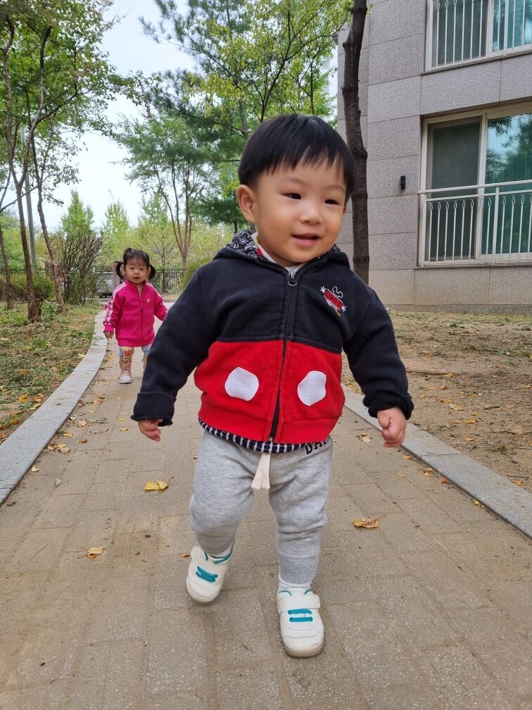 미키마우스 재킷을 입은 아기 지호가 가을 낙엽이 깔린 산책로를 걷고 있다. 환하게 웃고 있으며, 뒤편에는 핑크색 옷을 입은 다른 아이가 걸어오고 있다.