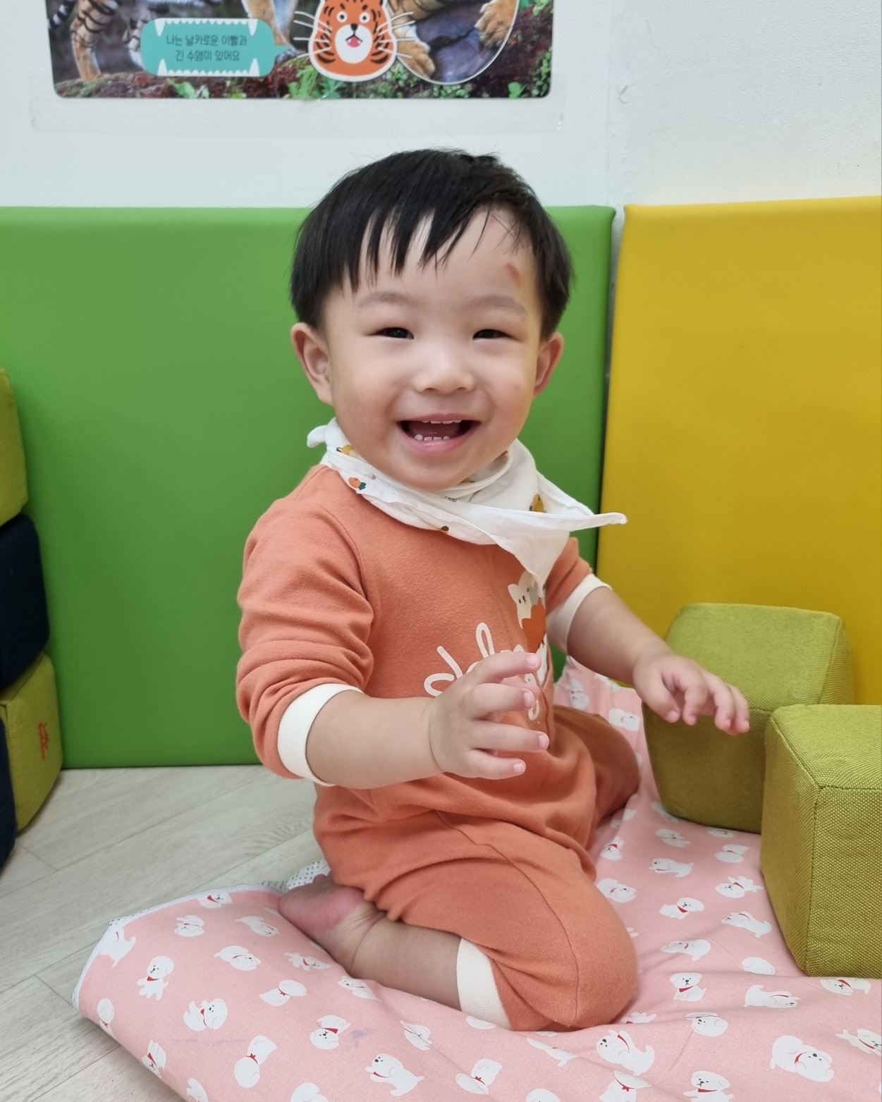 실내 놀이 공간으로 보이는 곳에서 오렌지색 상하복을 입은 아기가 분홍색 매트 위에 앉아 활짝 웃고 있다. 이마에는 작은 상처가 보이지만 해맑게 웃는 모습이 사랑스럽다. 배경에는 초록색과 노란색의 스펀지 블록들이 보인다.