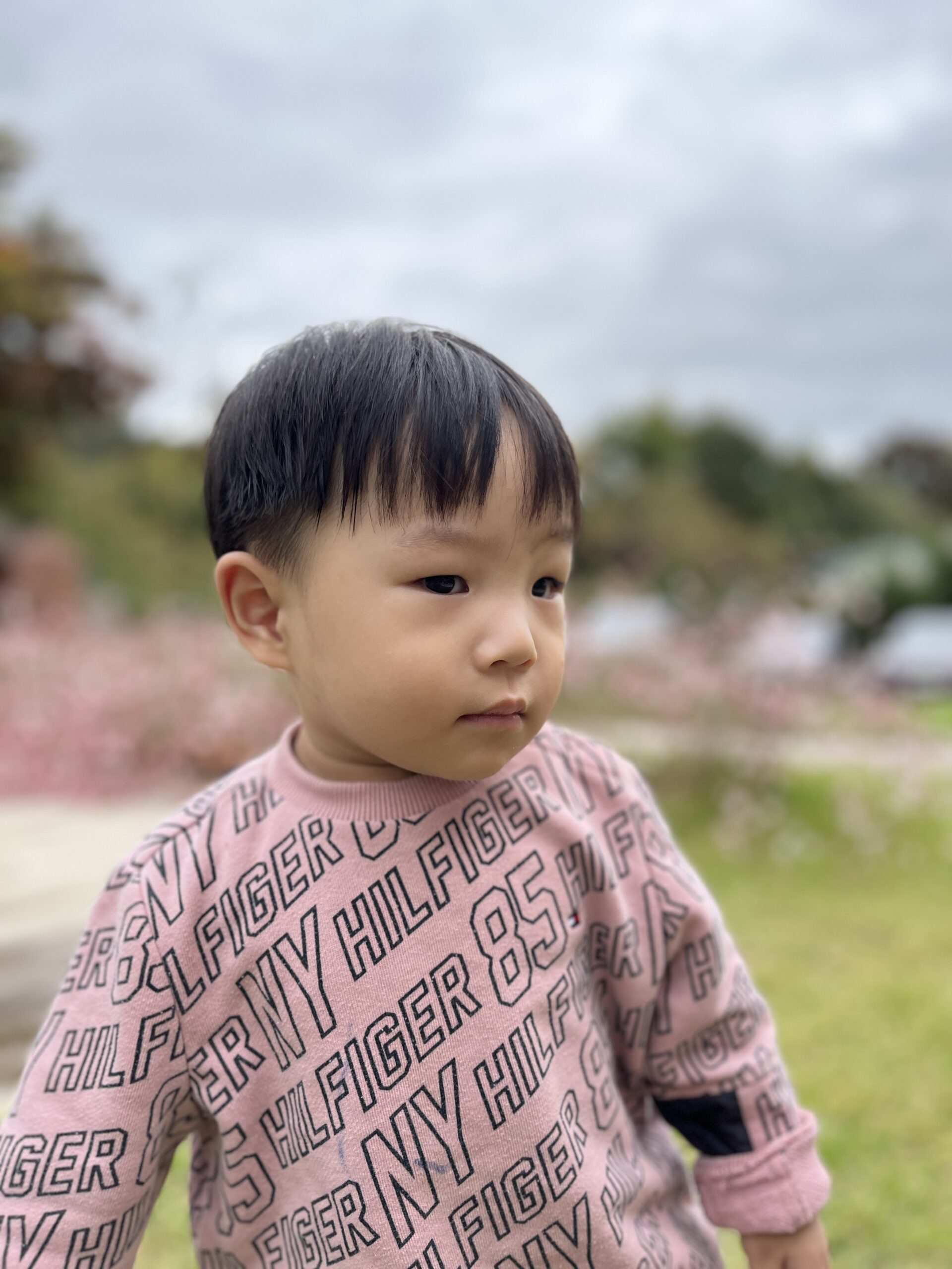야외 공원 같은 곳에서 분홍색 글씨가 인쇄된 스웨터와 베이지색 바지를 입은 아기가 잔디밭에 서서 왼쪽을 진지하게 바라보고 있다. 흐린 하늘과 흐릿한 핑크빛 꽃들이 배경으로 보인다.