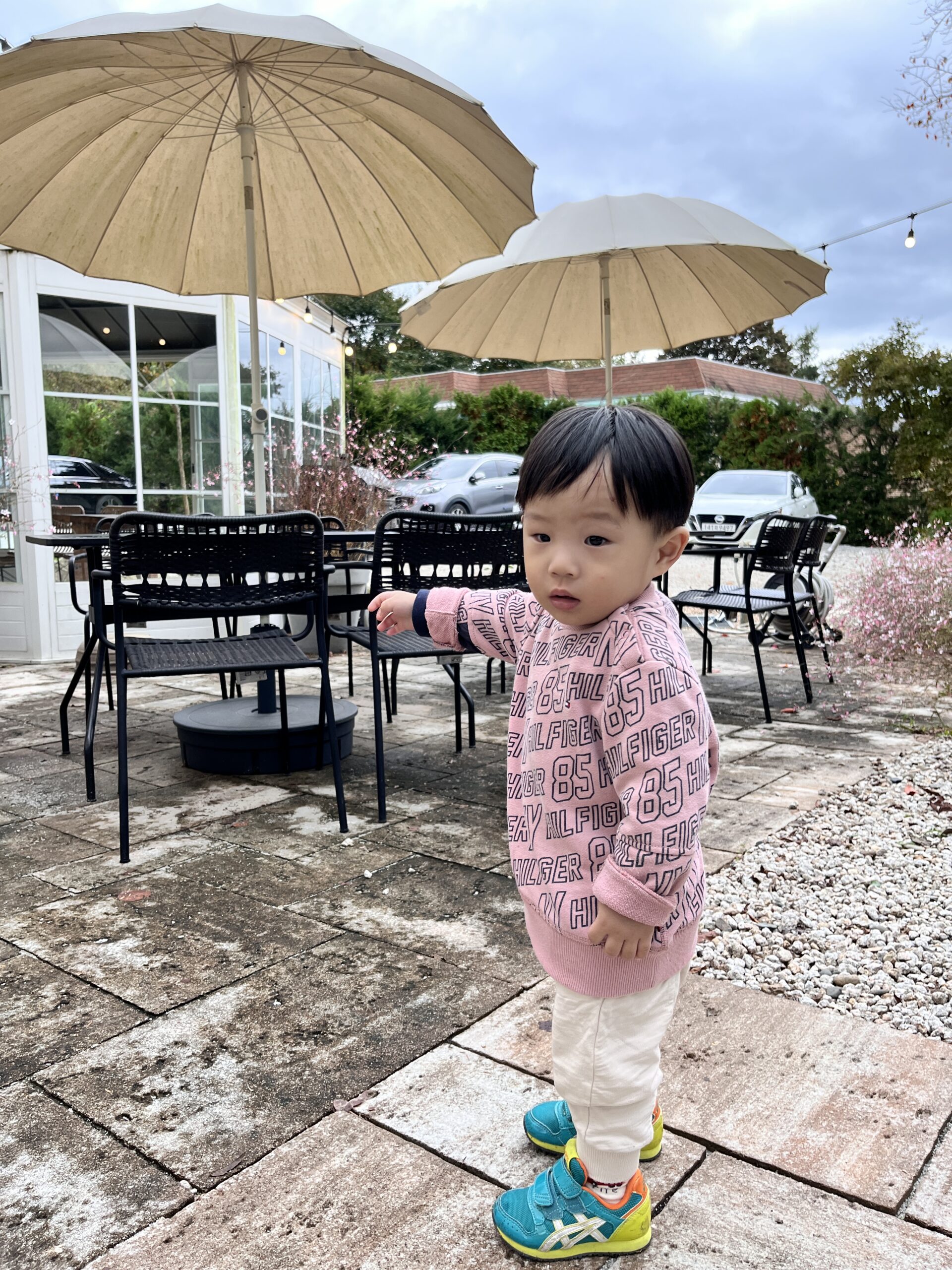 야외 카페 테라스로 보이는 곳에서 분홍색 스웨터와 베이지색 바지, 청록색 신발을 신은 아기가 서서 왼손을 들어 어딘가를 가리키고 있다. 뒤로는 야외 테이블과 의자, 파라솔, 그리고 차량들이 보인다. 바닥은 돌과 타일이 섞여 있다.