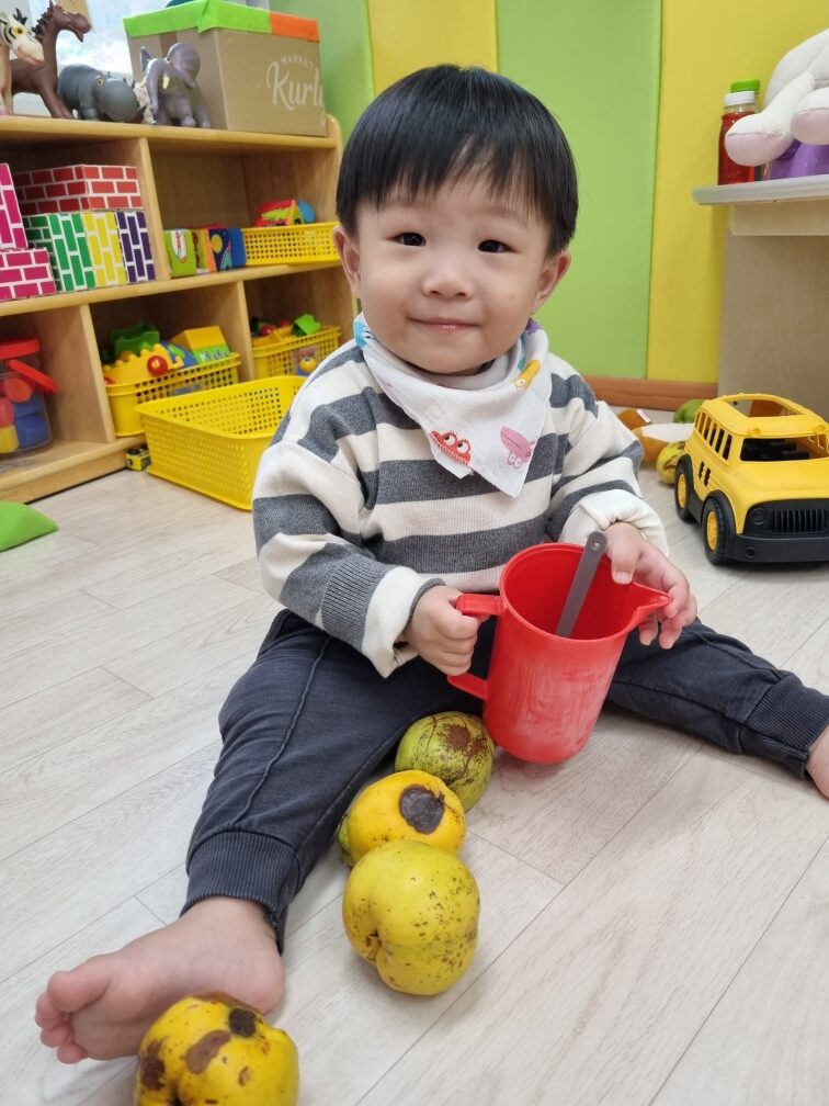 밝은 노란색과 회색 줄무늬 상의를 입고 맨발로 앉아있는 지호가 바닥에 놓인 노란색 과일들 옆에서 빨간색 컵과 숟가락을 들고 환하게 웃고 있다. 뒤쪽에는 장난감 선반이 보인다.