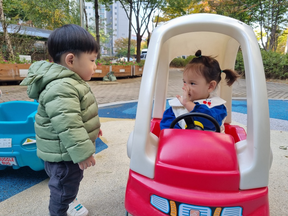 야외 놀이터에서 초록색 패딩을 입은 지호가 빨간색 장난감 자동차에 앉아있는 여자아이와 마주 보고 서 있다. 주변에는 나무와 건물들이 보인다.
