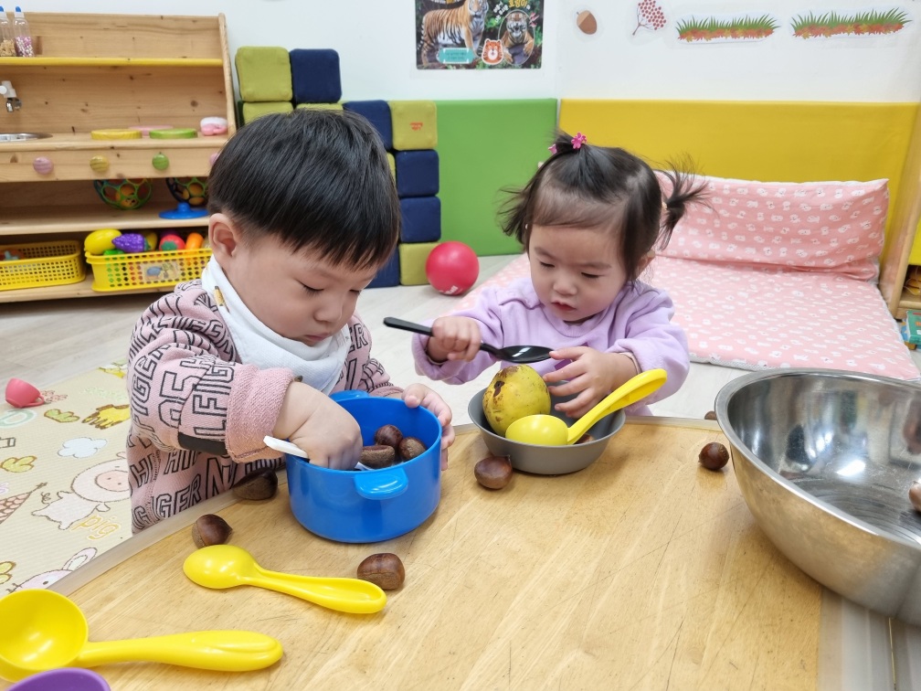 어린이집으로 보이는 실내 공간에서 지호가 분홍색 상의를 입고 친구와 함께 테이블에 앉아 밤과 노란색 과일을 이용한 놀이를 하고 있다. 지호는 파란색 그릇에 밤을 담고 있다.