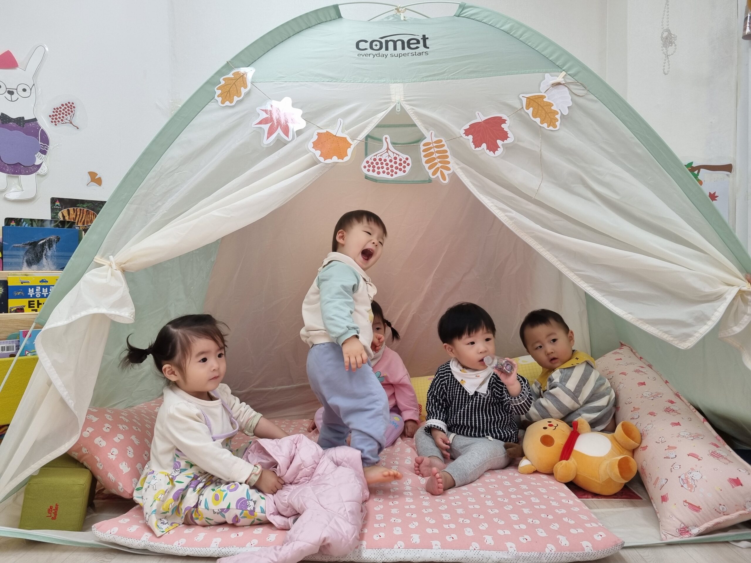 실내에 설치된 연두색 아기 텐트 안에 5명의 아기들이 모여 놀고 있다. 텐트 입구에는 가을 나뭇잎 모양의 장식들이 걸려있다. 한 아기는 텐트 안에서 벌떡 일어나 환하게 웃고 있고, 다른 아기들은 앉아서 인형이나 친구를 바라보고 있다.