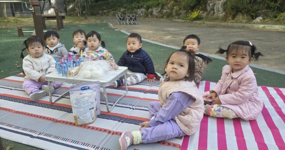푸른 잔디밭에 깔린 두 개의 담요 위에 7명의 아기들이 옹기종기 앉아있다. 중앙에는 작은 접이식 테이블이 있고 그 위에는 간식 봉투가 놓여있다. 아기들은 각자 다른 색깔의 가을옷을 입고 카메라를 바라보고 있다. 뒤로는 아파트 건물과 나무들이 보인다.