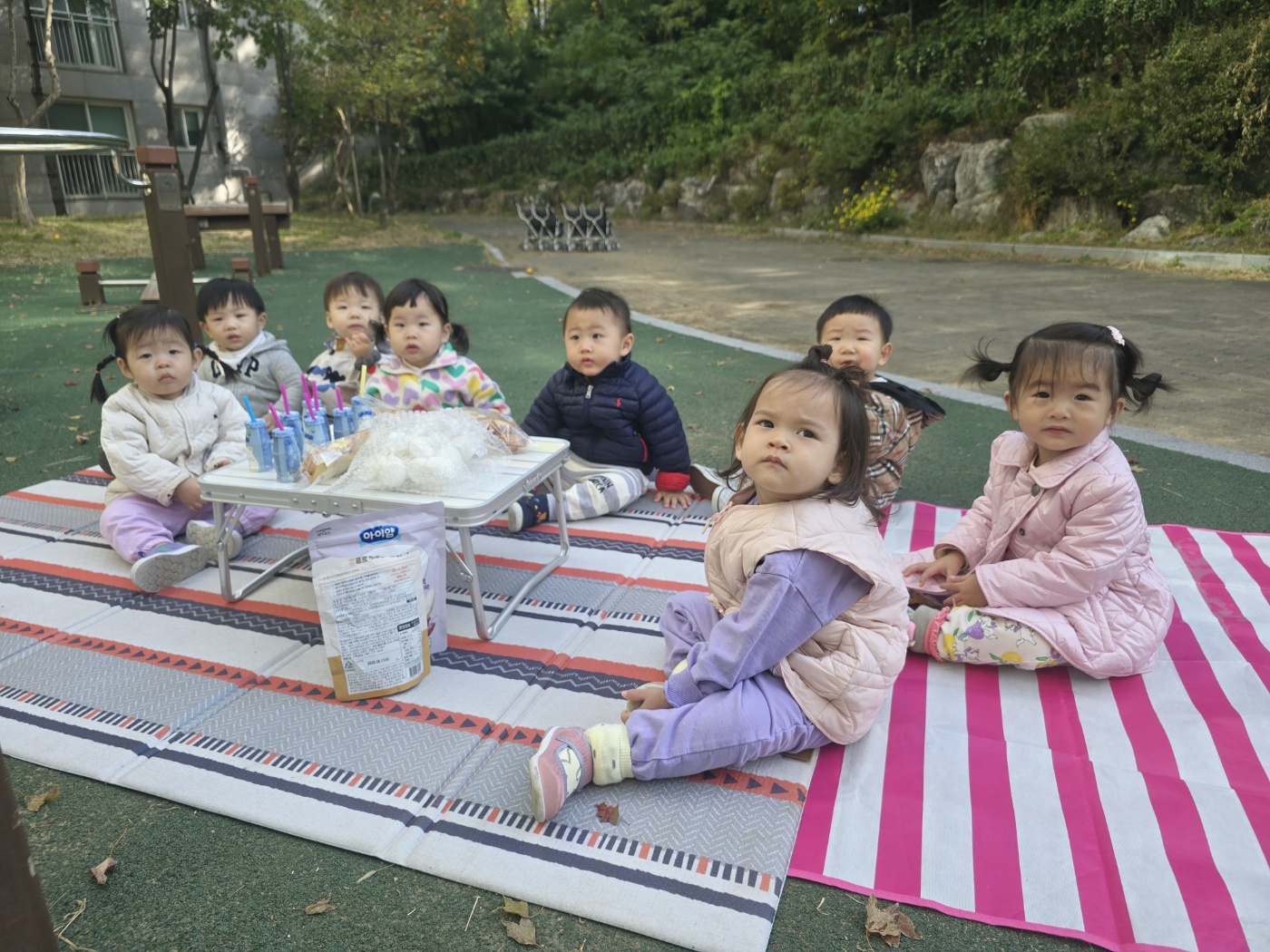 푸른 잔디밭에 깔린 두 개의 담요 위에 7명의 아기들이 옹기종기 앉아있다. 중앙에는 작은 접이식 테이블이 있고 그 위에는 간식 봉투가 놓여있다. 아기들은 각자 다른 색깔의 가을옷을 입고 카메라를 바라보고 있다. 뒤로는 아파트 건물과 나무들이 보인다.