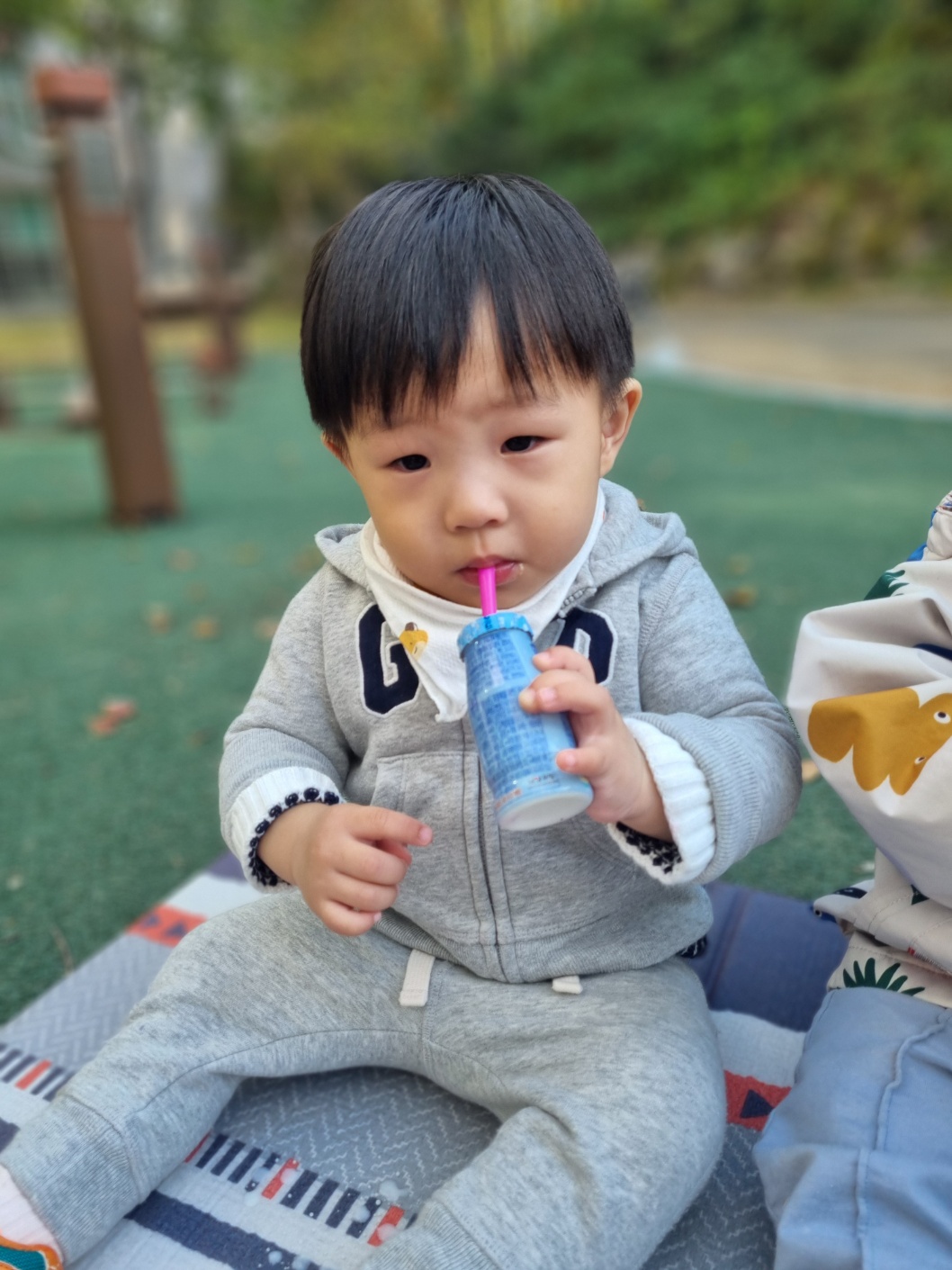 회색 GAP 후드티와 회색 트레이닝 바지를 입은 지호가 줄무늬 돗자리에 앉아 분홍색 빨대가 꽂힌 파란색 음료수를 마시고 있다. 뒤에는 놀이터 시설이 흐릿하게 보인다.