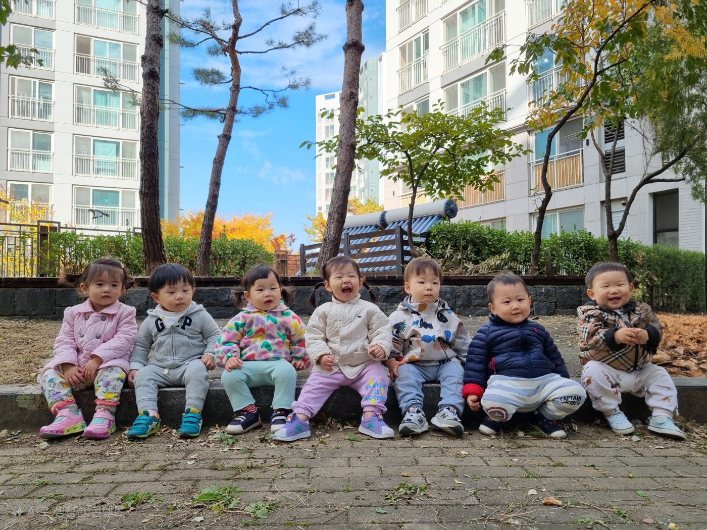 야외에서 7명의 아기들이 낮은 돌담 위에 나란히 앉아 카메라를 보고 웃고 있다. 중앙에 있는 아기는 입을 크게 벌리고 환하게 웃고 있으며, 다른 아기들도 미소를 짓고 있다. 뒤로는 아파트 건물과 노랗게 물든 가을 나무들이 보인다.