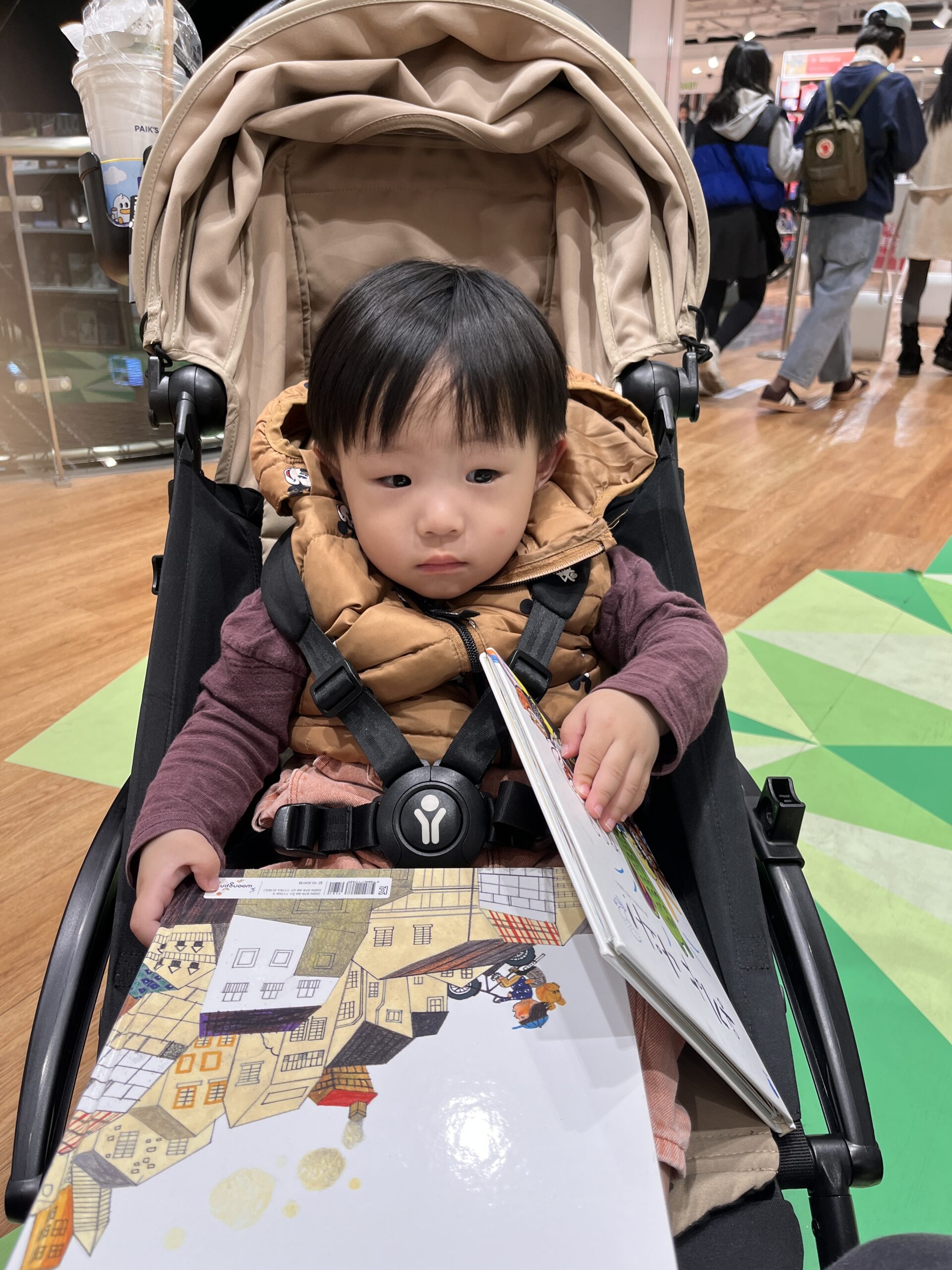 베이지색 유모차에 앉아있는 지호가 갈색 패딩 조끼를 입고 그림책을 펼쳐 보고 있다. 주변은 흐릿하게 처리된 실내 공간이며 사람들이 지나다니는 모습이 보인다.