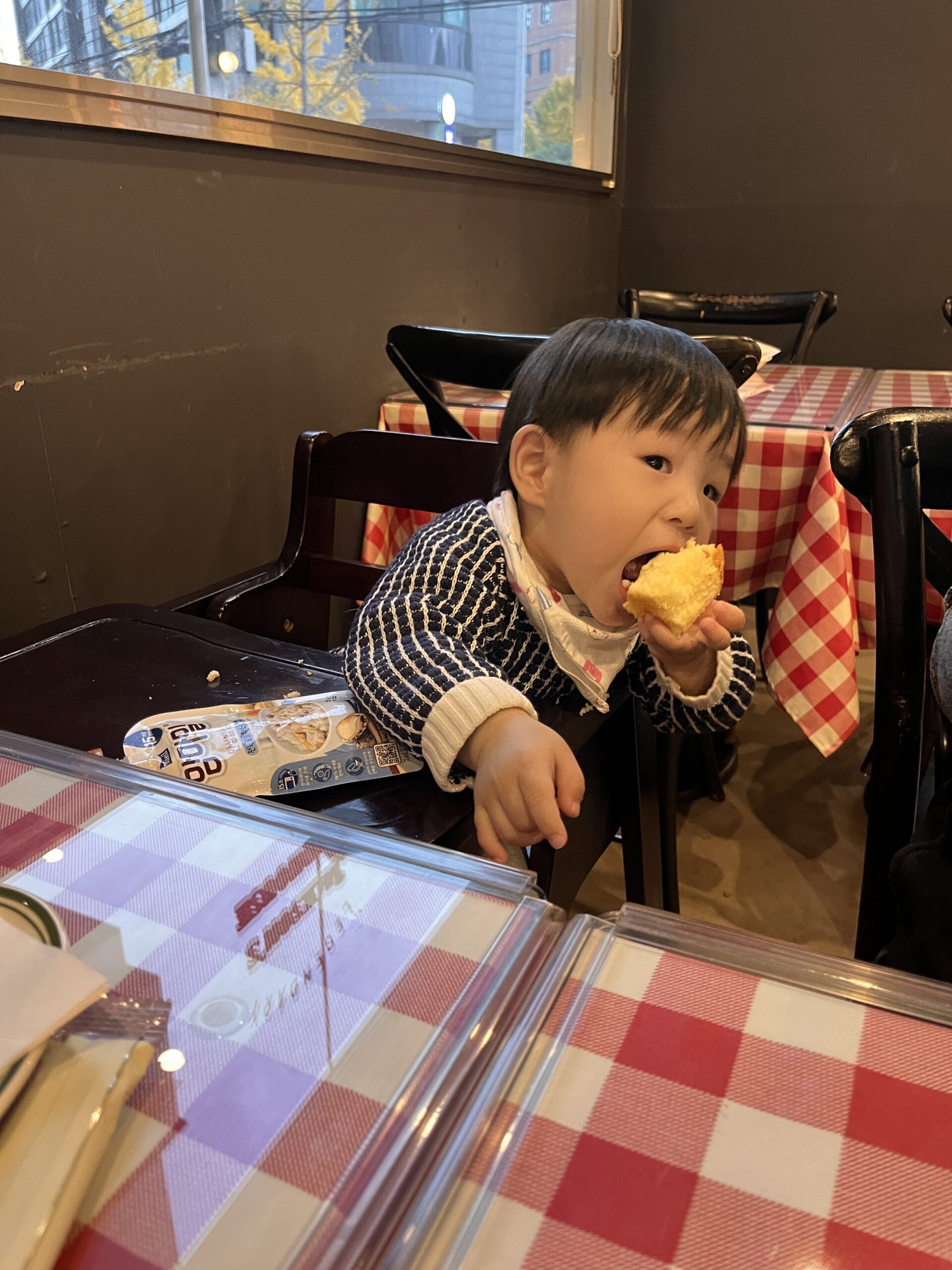 빨간색과 흰색 체크무늬 식탁보가 깔린 레스토랑 테이블 앞에 아기의자에 앉은 지호가 커다란 빵 조각을 두 손으로 잡고 입에 넣으며 맛있게 먹고 있다. 지호는 스트라이프 무늬 스웨터를 입고 있다.