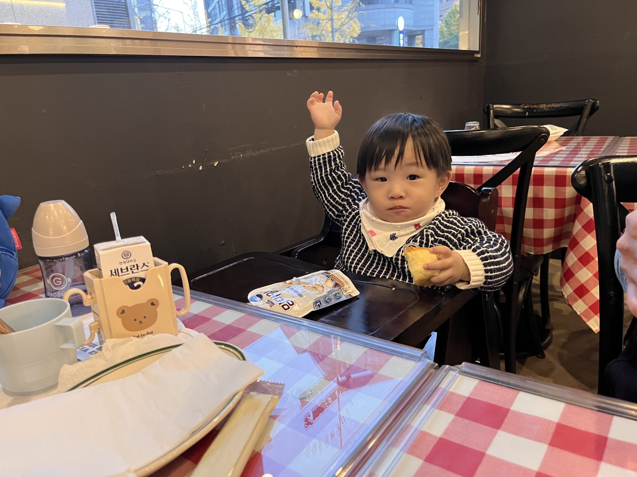 레스토랑 아기의자에 앉은 지호가 빵 조각을 한 손에 들고 다른 한 손을 번쩍 들고 카메라를 응시하고 있다. 테이블 위에는 아기용 물병과 우유팩, 식기가 놓여있다. 지호는 파란색 스트라이프 스웨터를 입고 있다.