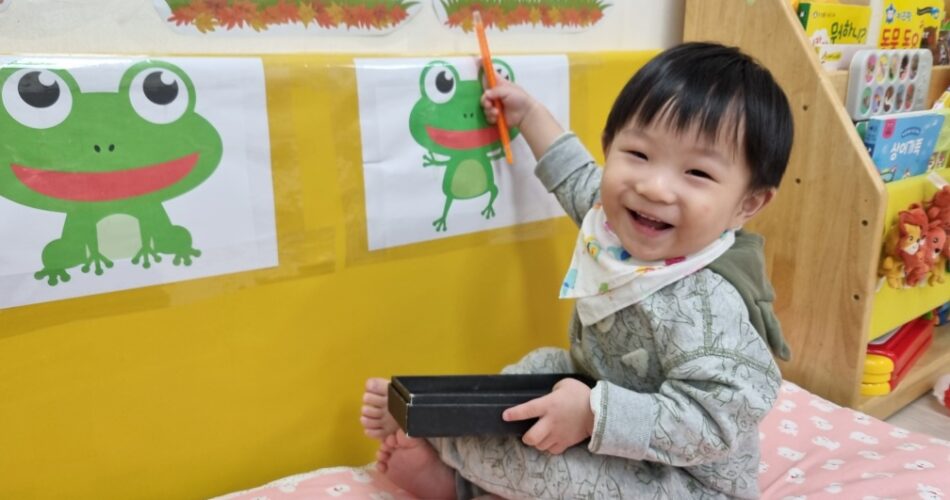 분홍색 매트 위에 앉아 활짝 웃는 지호가 개구리 그림을 향해 주황색 연필을 들고 있다. 배경에는 노란 벽과 책장이 보인다.