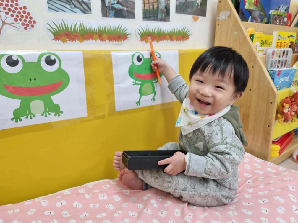 분홍색 매트 위에 앉아 활짝 웃는 지호가 개구리 그림을 향해 주황색 연필을 들고 있다. 배경에는 노란 벽과 책장이 보인다.