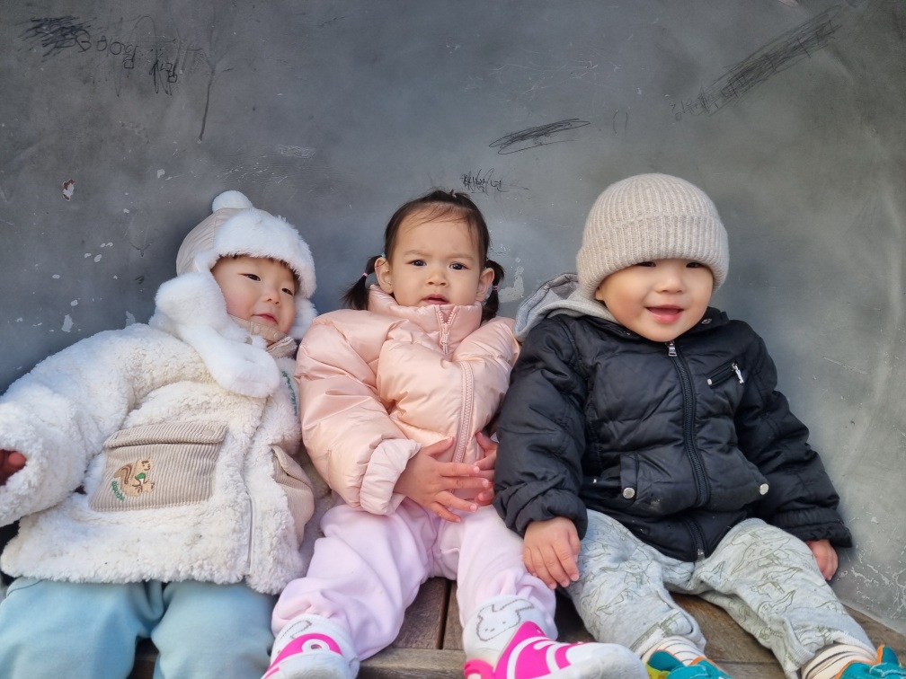 회색 벽을 배경으로 나란히 앉아있는 세 아이. 오른쪽의 지호는 검은색 패딩과 털모자를 쓰고 환하게 웃고 있고, 가운데 여자아이는 분홍색 패딩을, 왼쪽 아이는 흰색 털 외투를 입고 있다.