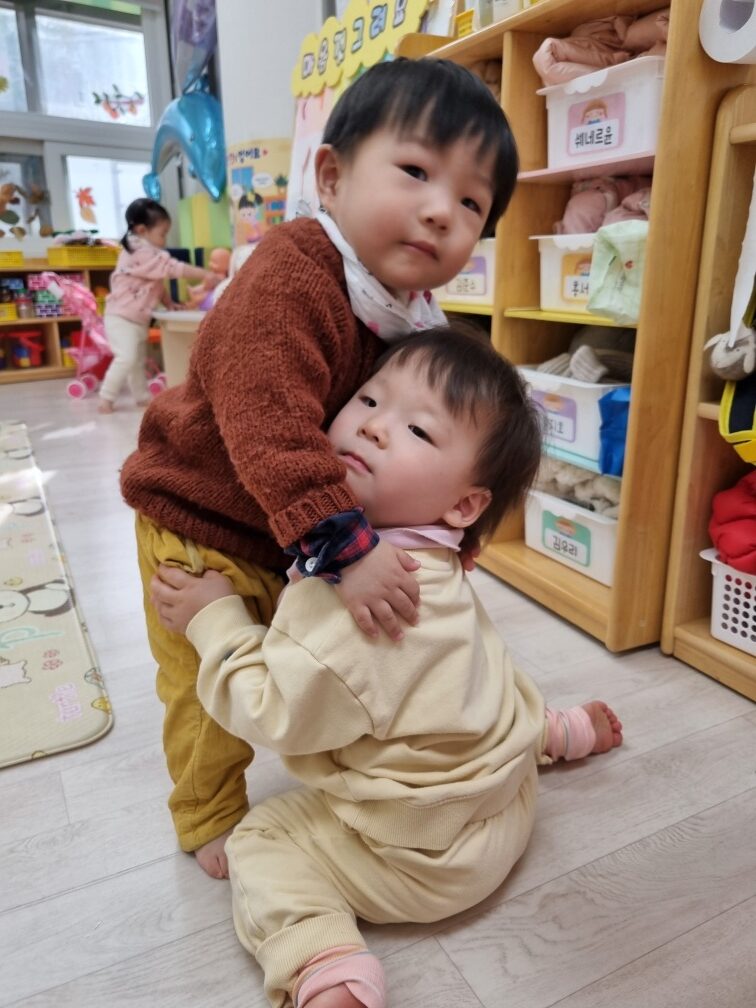 갈색 스웨터를 입은 지호가 바닥에 앉아있는 친구를 다정하게 안아주고 있다. 두 아이는 어린이집 교실로 보이는 공간에 있다.