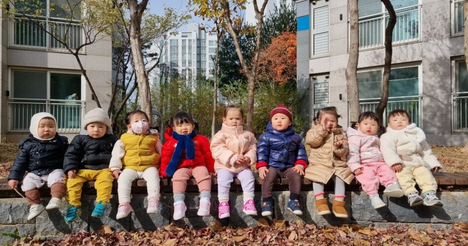 알록달록한 낙엽이 흩뿌려진 보도블럭 옆 낮은 돌담 위에 9명의 아기들이 겨울 외투를 입고 옹기종기 앉아 밝은 햇살을 받고 있는 모습입니다. 아파트 단지 내의 야외 공간으로 보이며, 배경에는 아파트 건물과 단풍이 든 나무들이 보입니다.