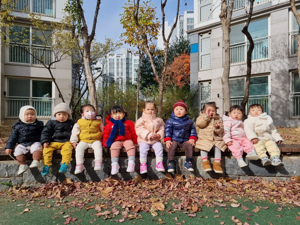 알록달록한 낙엽이 흩뿌려진 보도블럭 옆 낮은 돌담 위에 9명의 아기들이 겨울 외투를 입고 옹기종기 앉아 밝은 햇살을 받고 있는 모습입니다. 아파트 단지 내의 야외 공간으로 보이며, 배경에는 아파트 건물과 단풍이 든 나무들이 보입니다.