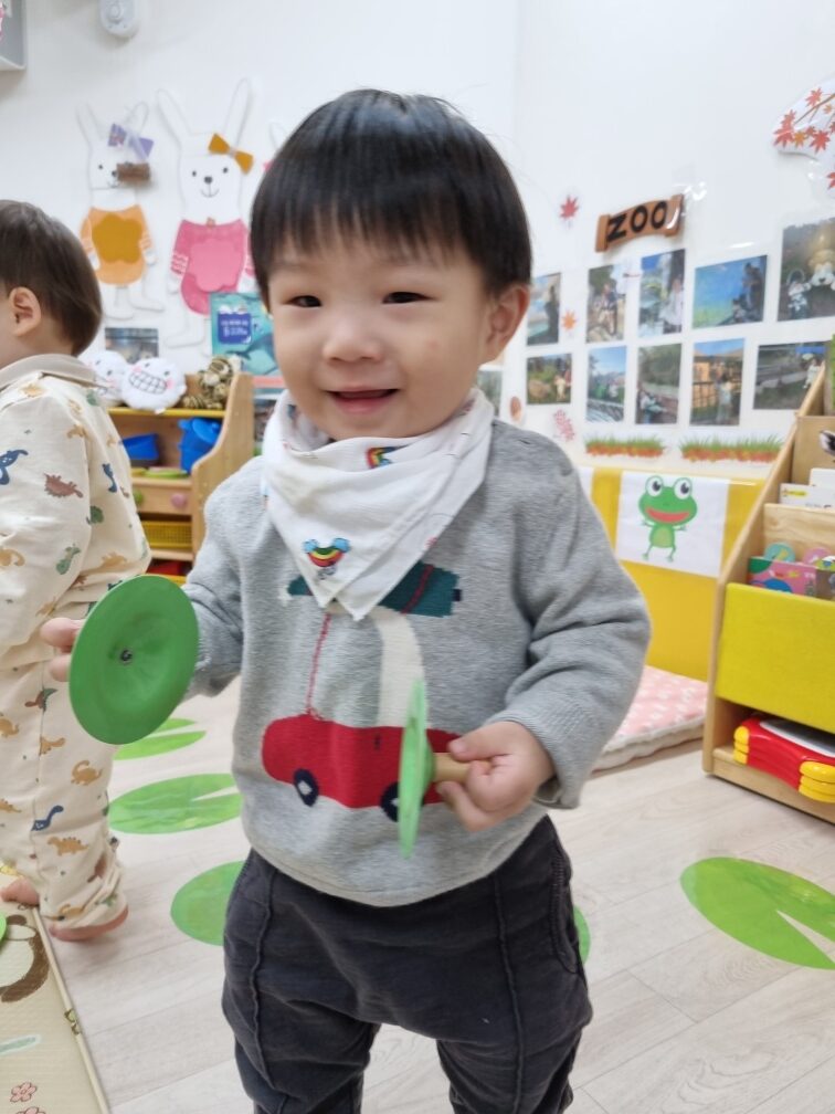 회색 상의를 입고 흰색 턱받이를 한 아이가 초록색 짝짝이 악기 두 개를 들고 환하게 웃으며 카메라를 바라보고 있다. 배경은 어린이집 교실처럼 보인다.