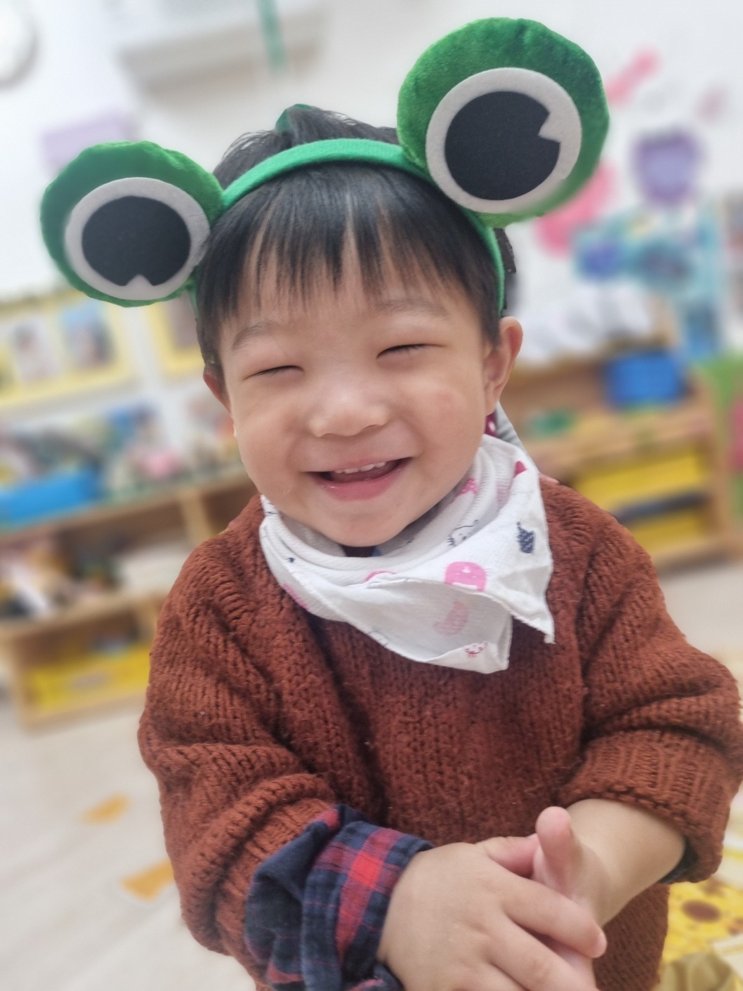 실내에서 브라운색 스웨터와 체크 셔츠를 입고 개구리 모양의 초록색 머리띠를 한 지호가 눈을 꼭 감고 해맑게 웃고 있는 클로즈업 사진입니다. 지호의 얼굴에는 행복한 미소가 가득합니다. 배경은 흐릿하게 처리되어 실내 교실임을 짐작할 수 있습니다.