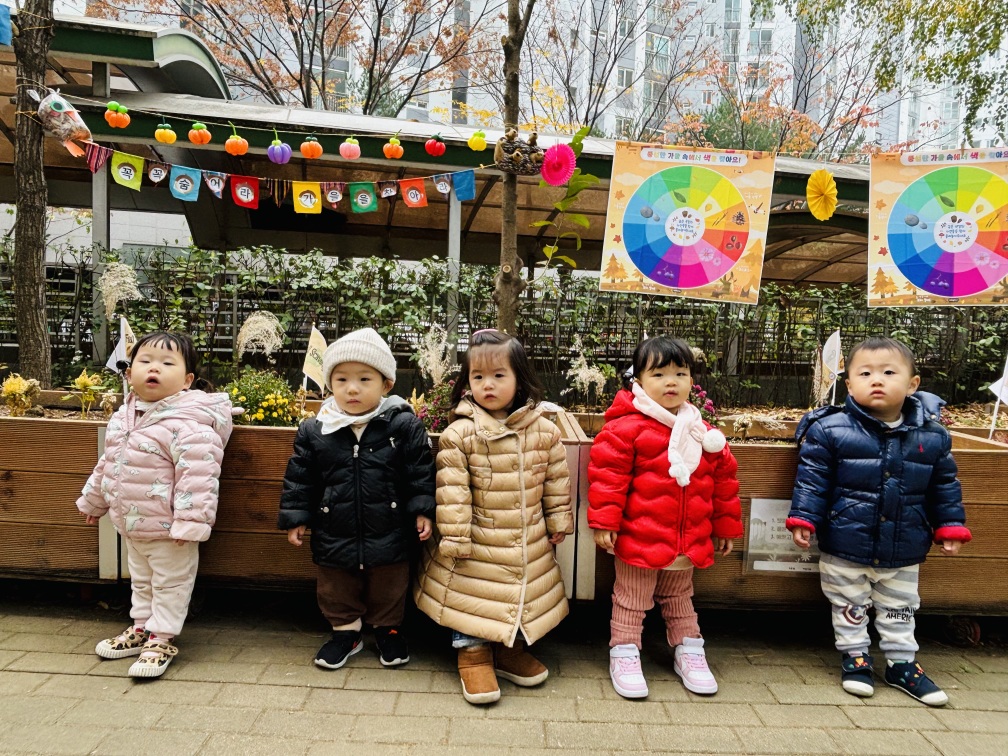 어린이집 야외 공간에서 5명의 아기들이 겨울 외투를 입고 일렬로 서서 카메라를 바라보고 있습니다. 아기들 뒤편으로는 가을 분위기의 장식과 색상환 교육 자료가 걸려 있습니다. 바닥에는 낙엽이 드문드문 보입니다.