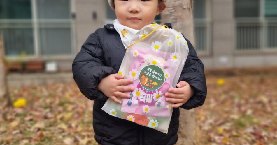 검은색 패딩과 베이지색 비니를 쓴 지호가 낙엽이 쌓인 나무 앞에서 미소 없이 정면을 응시하며 "생일 축하해" 문구가 적힌 선물 꾸러미를 두 손으로 소중히 들고 서 있다.