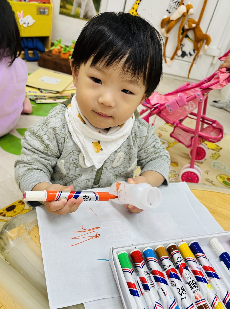 흰색 손수건 턱받이를 한 아이가 낮은 테이블에 앉아 종이에 주황색 마커로 그림을 그리고 있다. 옆에는 다양한 색깔의 마커들이 놓여 있다.