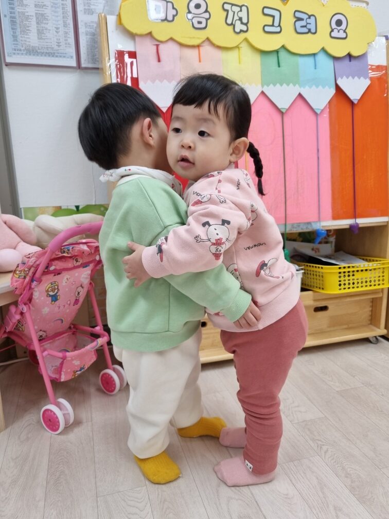 연두색 상의를 입은 지호가 실내에서 분홍색 옷을 입은 친구를 뒤에서 끌어안고 있다. 뒤쪽 벽에는 '마음껏 그려요'라는 문구가 보인다.