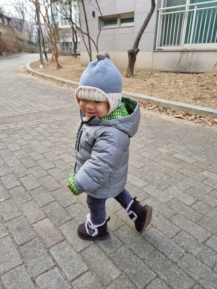 회색 패딩과 파란색 비니를 쓴 지호가 보도블록 길을 따라 걷고 있다. 옆모습이 보이며, 건물과 나무가 배경에 흐릿하게 보인다.
