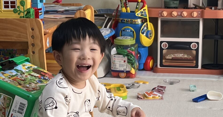 따스한 햇살이 드는 방에서 판다 잠옷을 입은 지호가 바닥에 앉아 알록달록 블록을 가지고 놀며 활짝 웃고 있다. 뒤쪽으로는 장난감 부엌과 인형의 집이 보인다.