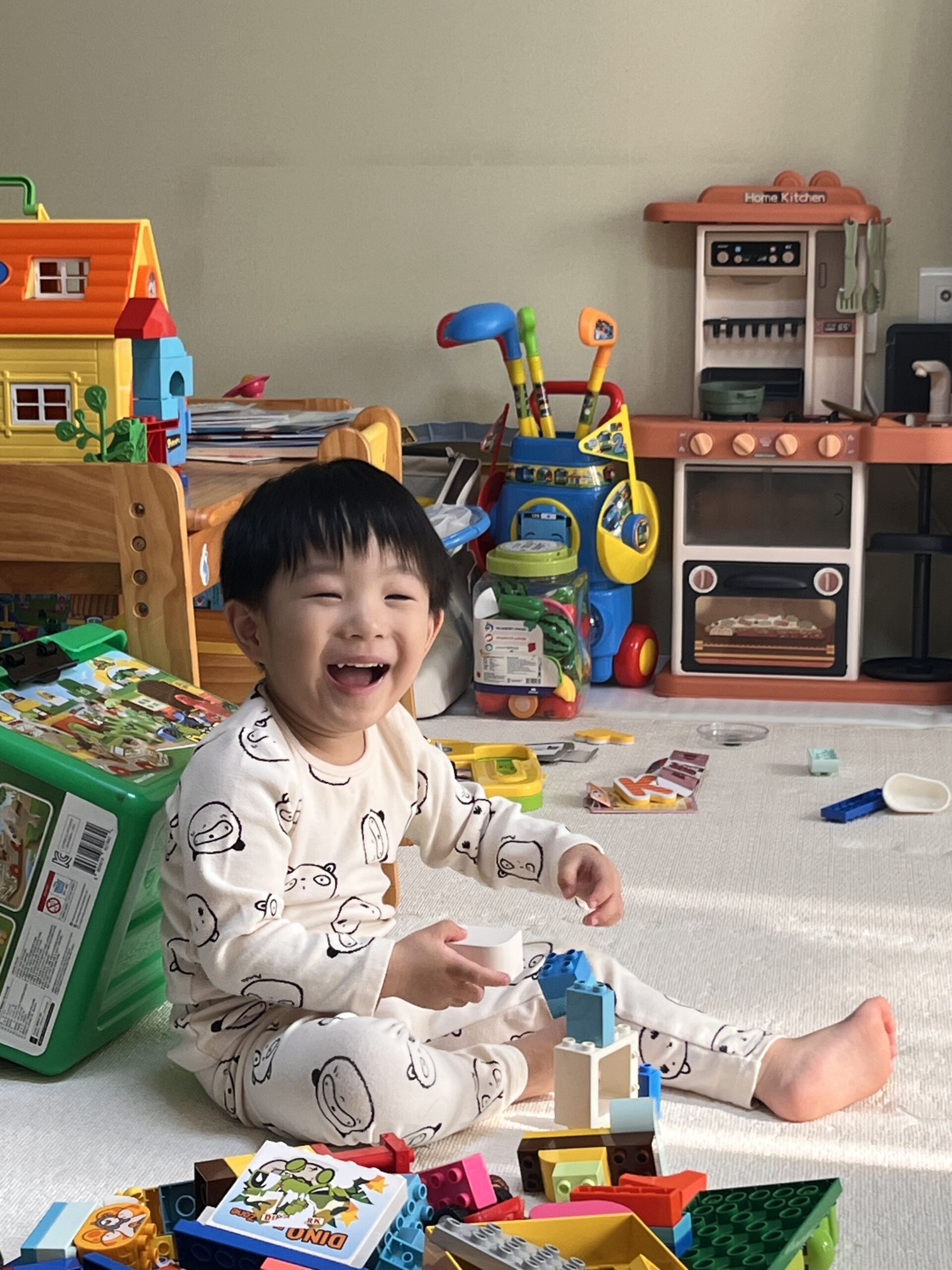 따스한 햇살이 드는 방에서 판다 잠옷을 입은 지호가 바닥에 앉아 알록달록 블록을 가지고 놀며 활짝 웃고 있다. 뒤쪽으로는 장난감 부엌과 인형의 집이 보인다.