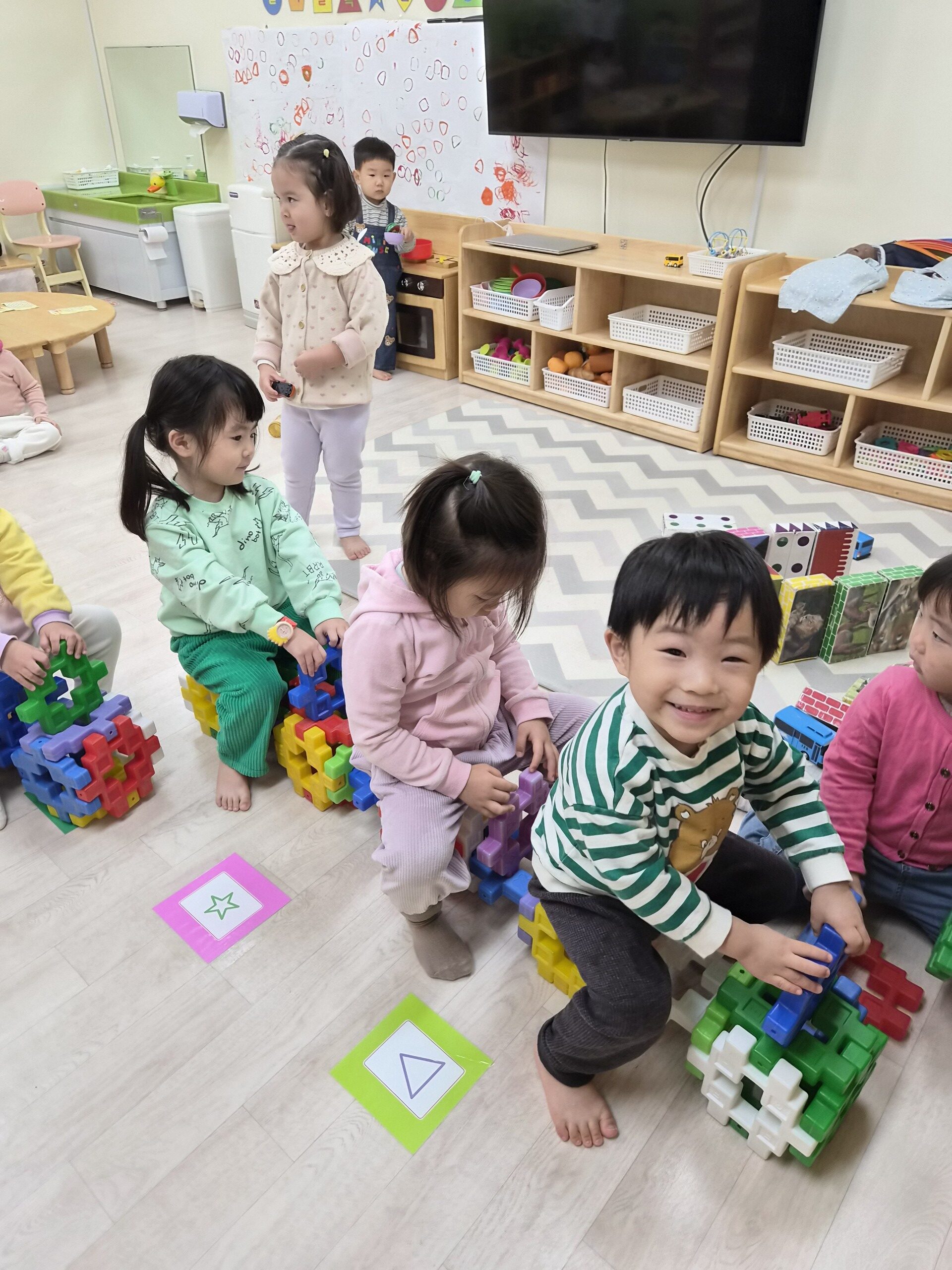 어린이집 교실에서 지호가 초록색 줄무늬 옷을 입고 다른 아이들과 함께 바닥에 앉아 커다란 블록으로 기차를 만들고 있다. 지호는 카메라를 보며 활짝 웃고 있다.