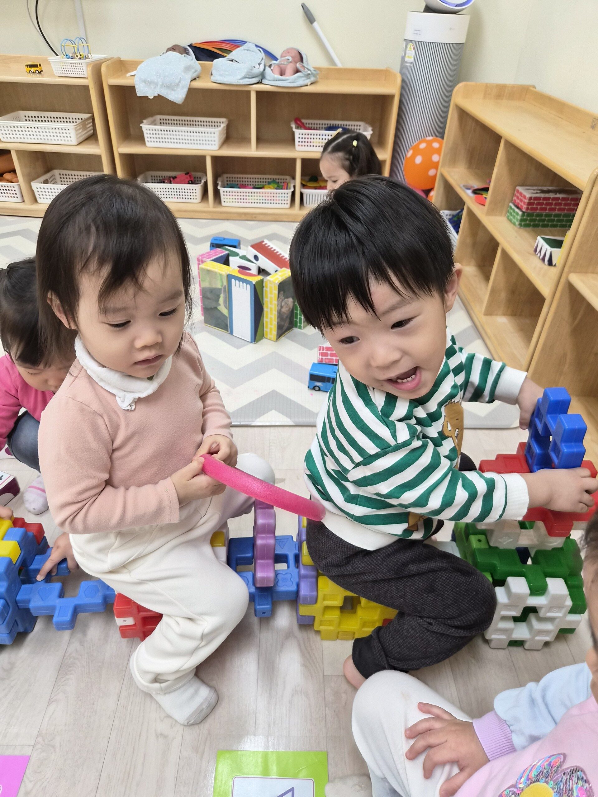 어린이집에서 지호가 초록색 줄무늬 옷을 입고 바닥에 앉아 옆에 앉은 여자아이와 함께 커다란 블록을 연결하며 진지하게 놀이에 집중하고 있다.
