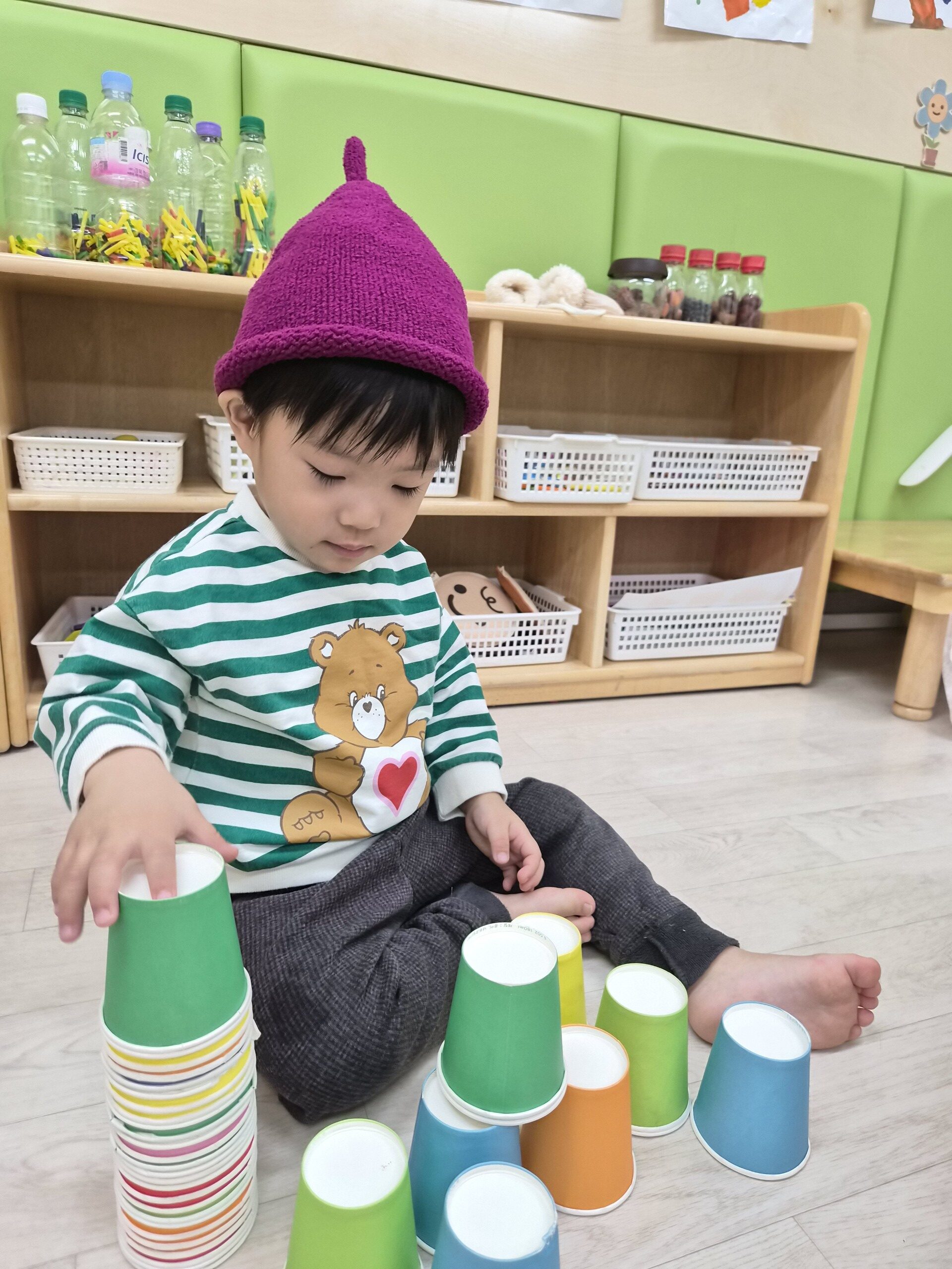 어린이집에서 지호가 초록색 줄무늬 옷과 보라색 털모자를 쓰고 바닥에 앉아 알록달록한 종이컵을 차곡차곡 쌓으며 놀이에 집중하고 있다.