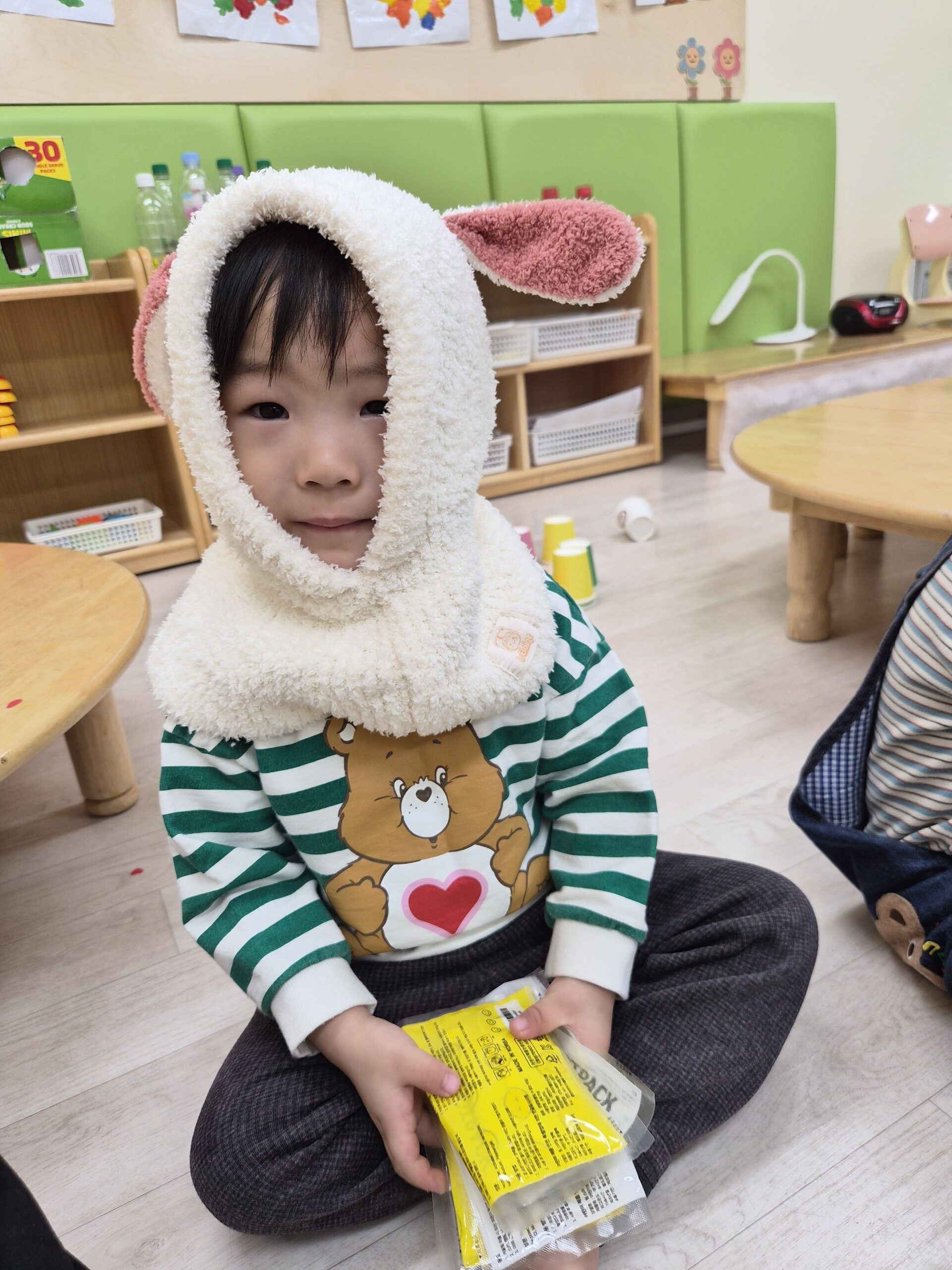 어린이집에서 지호가 초록색 줄무늬 옷에 하얀색 토끼 귀 모양 털모자를 쓰고 작은 노란색 봉투를 들고 카메라를 향해 온화하게 미소 짓고 있다.