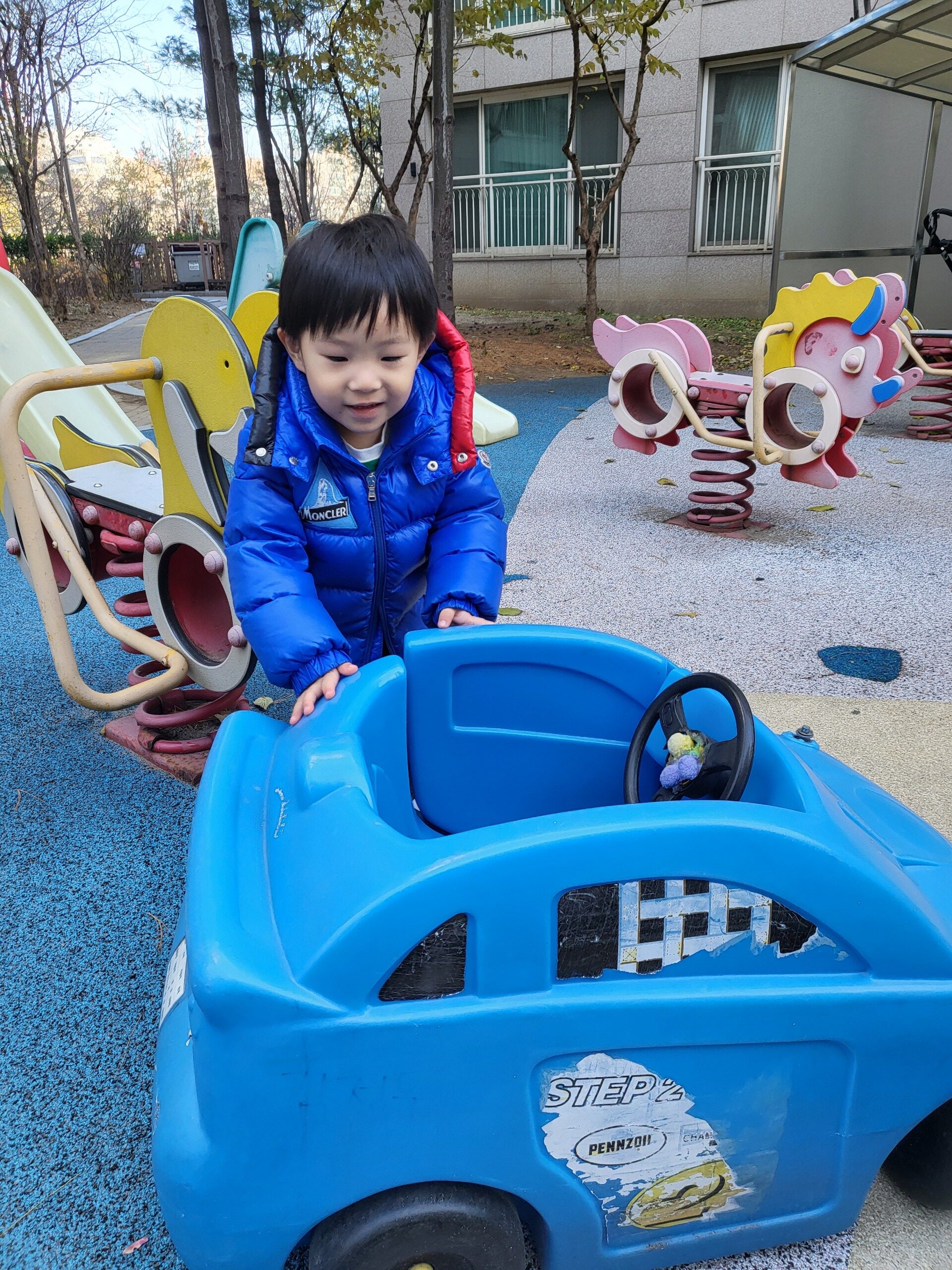 놀이터에서 지호가 파란색 패딩을 입고 파란색 장난감 자동차 옆에 서서 아래를 내려다보고 있다. 뒤로는 빨간색과 노란색의 흔들 놀이기구들이 보인다.