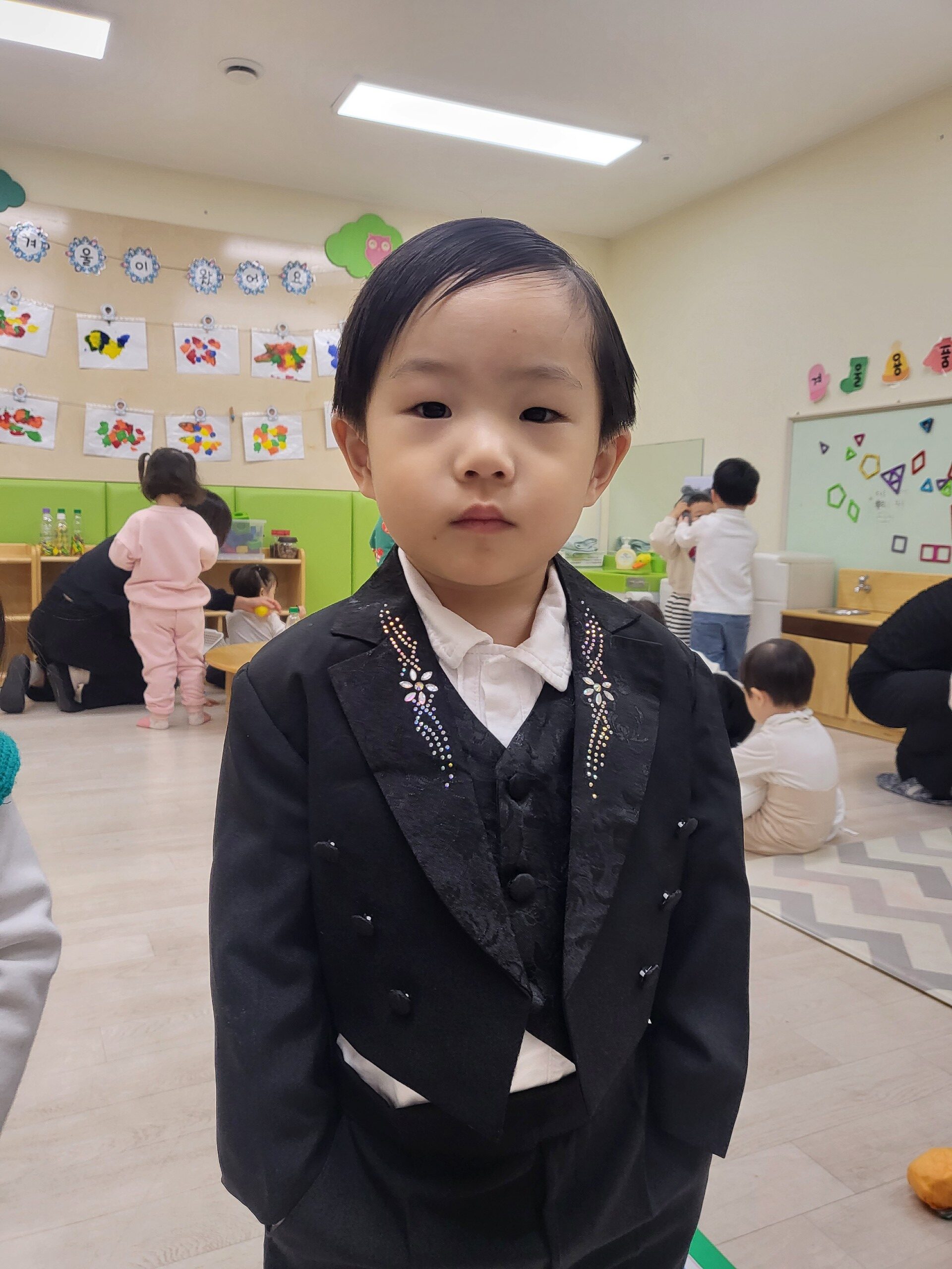 실내 보육 공간에서 지호가 검은색 턱시도와 흰색 셔츠를 입고 정면을 응시하고 있다. 그의 뒤편으로는 다른 아이들과 장식물들이 보인다.