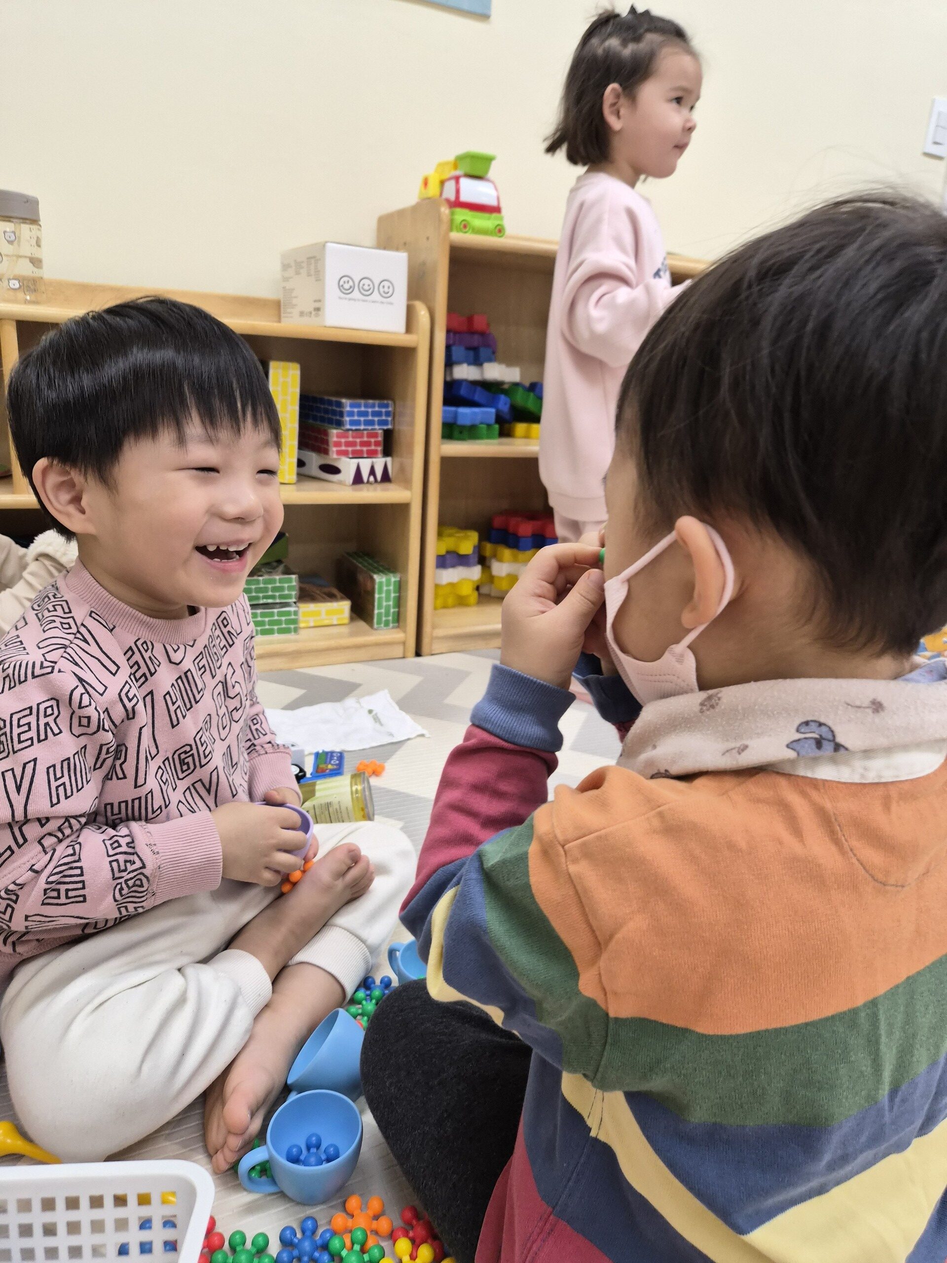 연한 분홍색 맨투맨 티셔츠를 입은 지호가 바닥에 앉아 친구와 마주 보며 블록 조각들로 놀고 있다. 지호는 활짝 웃으며 친구와 소통하고 있고, 뒤로는 다른 아이들이 보인다.