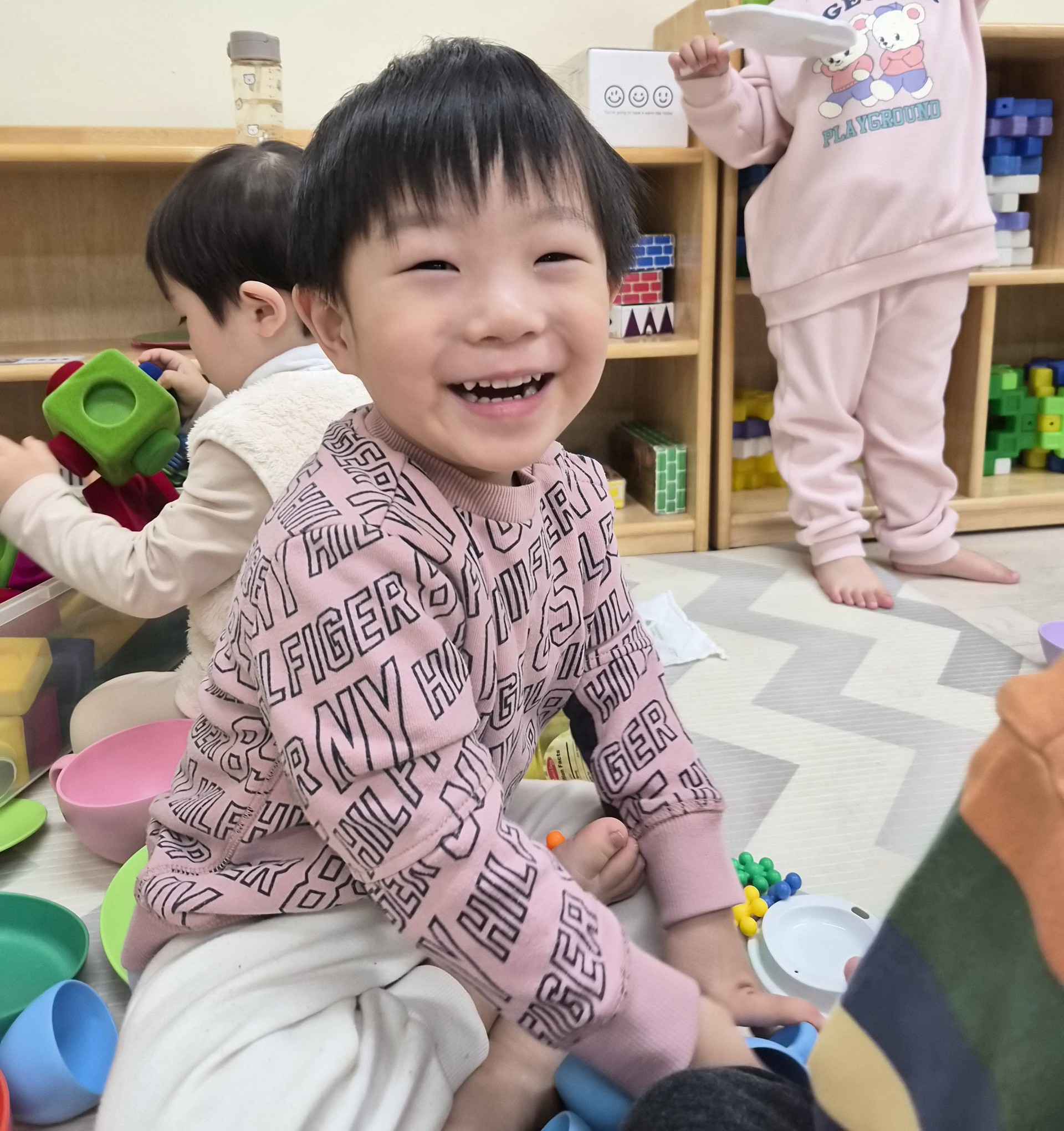 지호가 분홍색 상의를 입고 맨발로 앉아 카메라를 보며 환하게 웃고 있다. 주변에는 알록달록한 플라스틱 그릇과 장난감들이 흩어져 있고, 뒤쪽에는 다른 아이들이 놀고 있다.