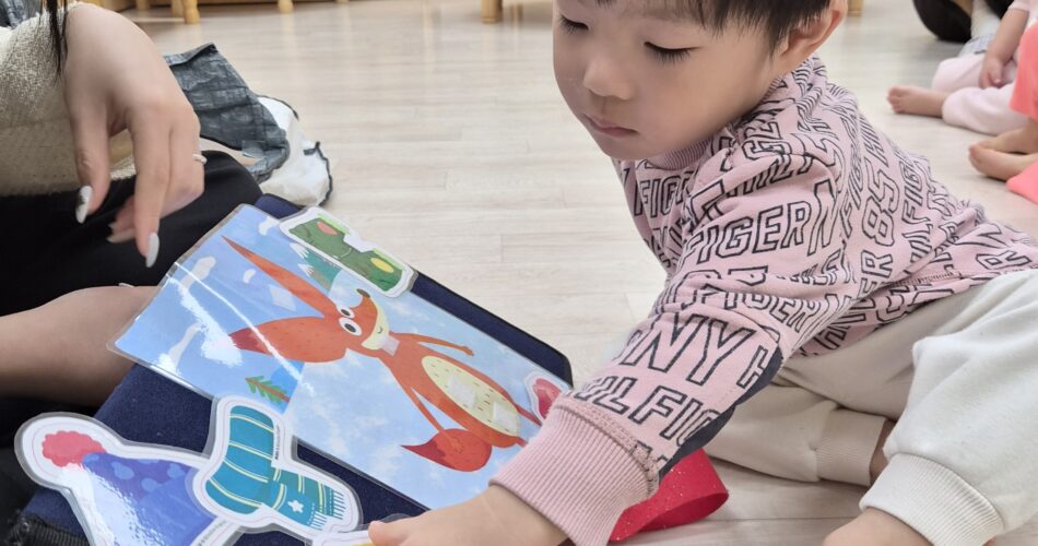 지호가 분홍색 상의를 입고 맨발로 앉아 펠트 보드에 있는 동물 모양 조각들을 손으로 만지며 집중하고 있다. 옆에는 어른의 손이 보이며, 뒤쪽 벽에는 크리스마스 장식이 걸려 있다.