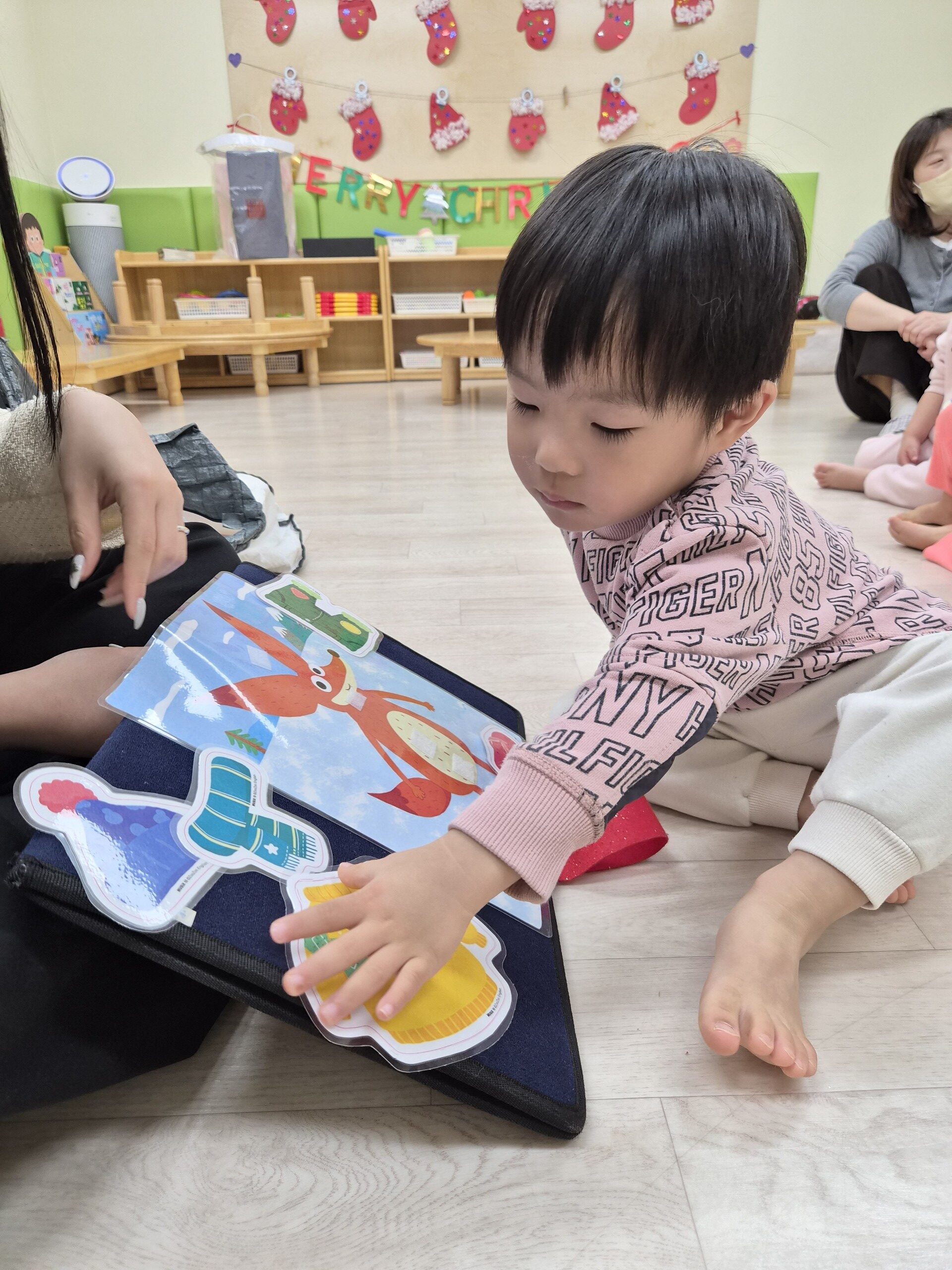 지호가 분홍색 상의를 입고 맨발로 앉아 펠트 보드에 있는 동물 모양 조각들을 손으로 만지며 집중하고 있다. 옆에는 어른의 손이 보이며, 뒤쪽 벽에는 크리스마스 장식이 걸려 있다.