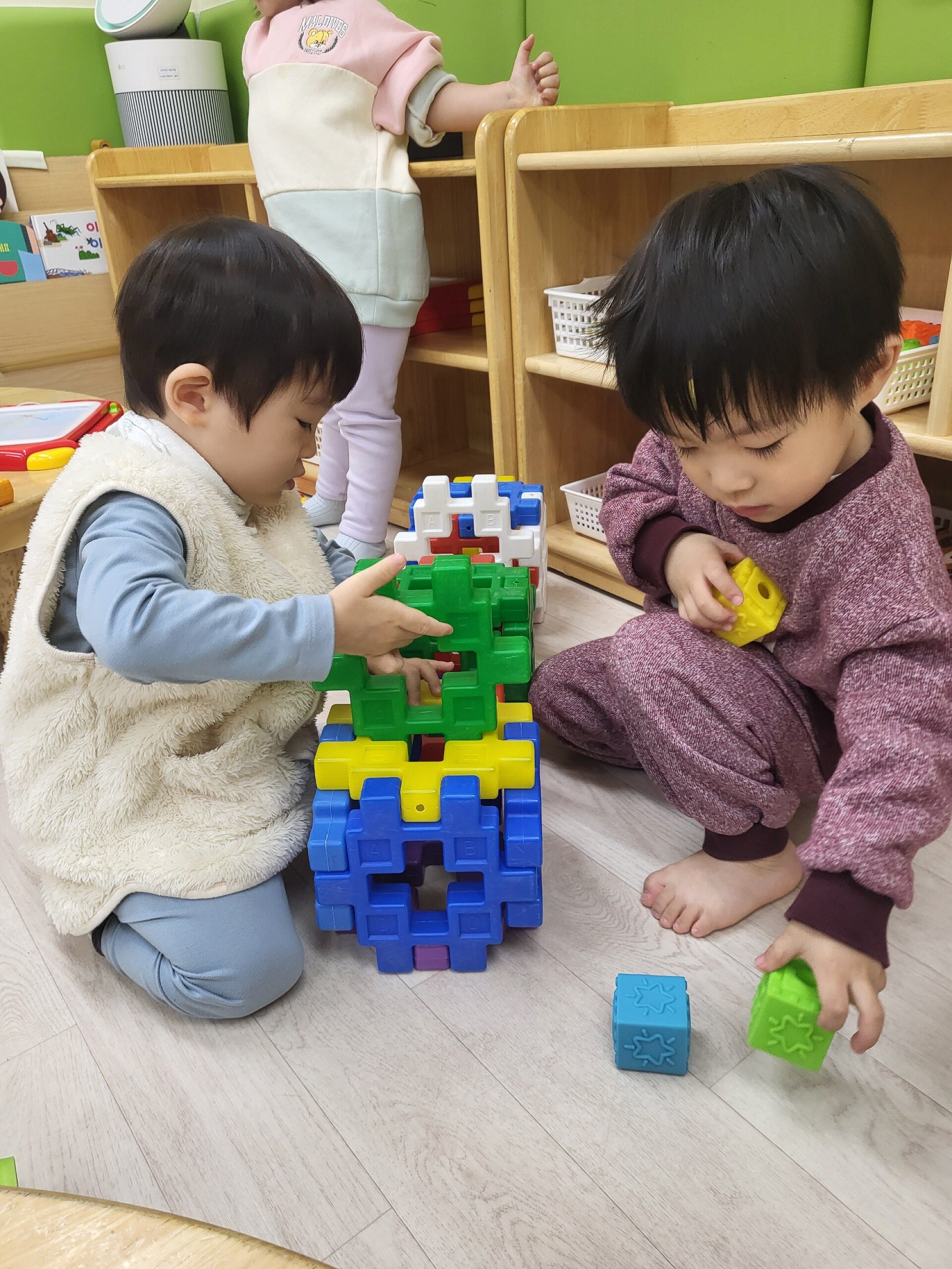 지호가 자주색 상하의를 입고 맨발로 앉아 옆 친구와 함께 알록달록한 블록으로 구조물을 만들고 있다. 지호는 녹색 블록을 들고 있으며, 친구는 이미 큰 블록 구조물을 만들고 있다.