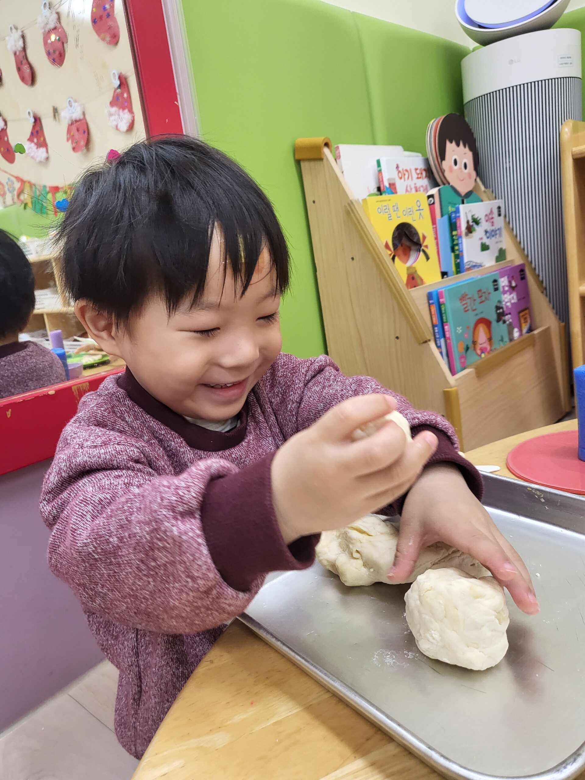 지호가 자주색 상의를 입고 작은 반죽 덩어리를 들고 환하게 웃고 있다. 앞에는 스테인리스 쟁반 위에 다른 반죽 덩어리들이 놓여 있으며, 배경에는 책장과 초록색 벽이 보인다.